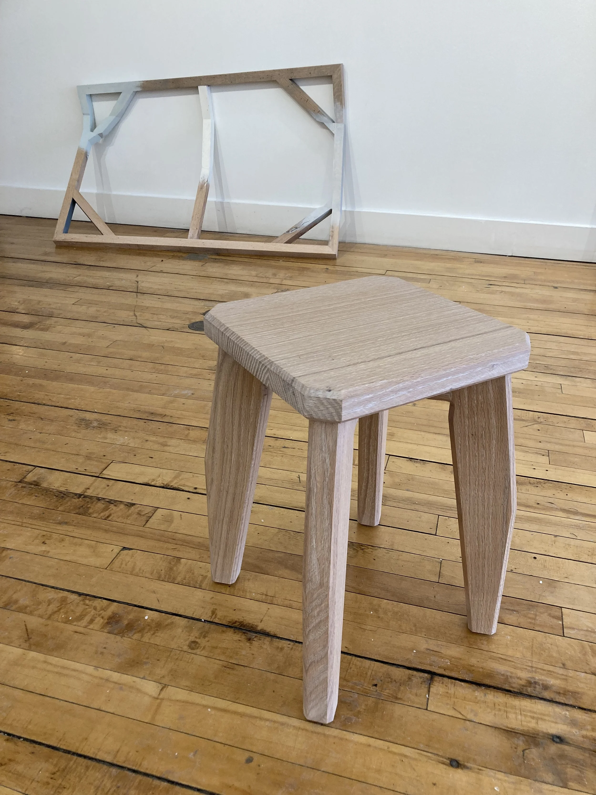 A small wooden stool with a square top on a wooden floor, with a wooden frame leaning against the white wall in the background.