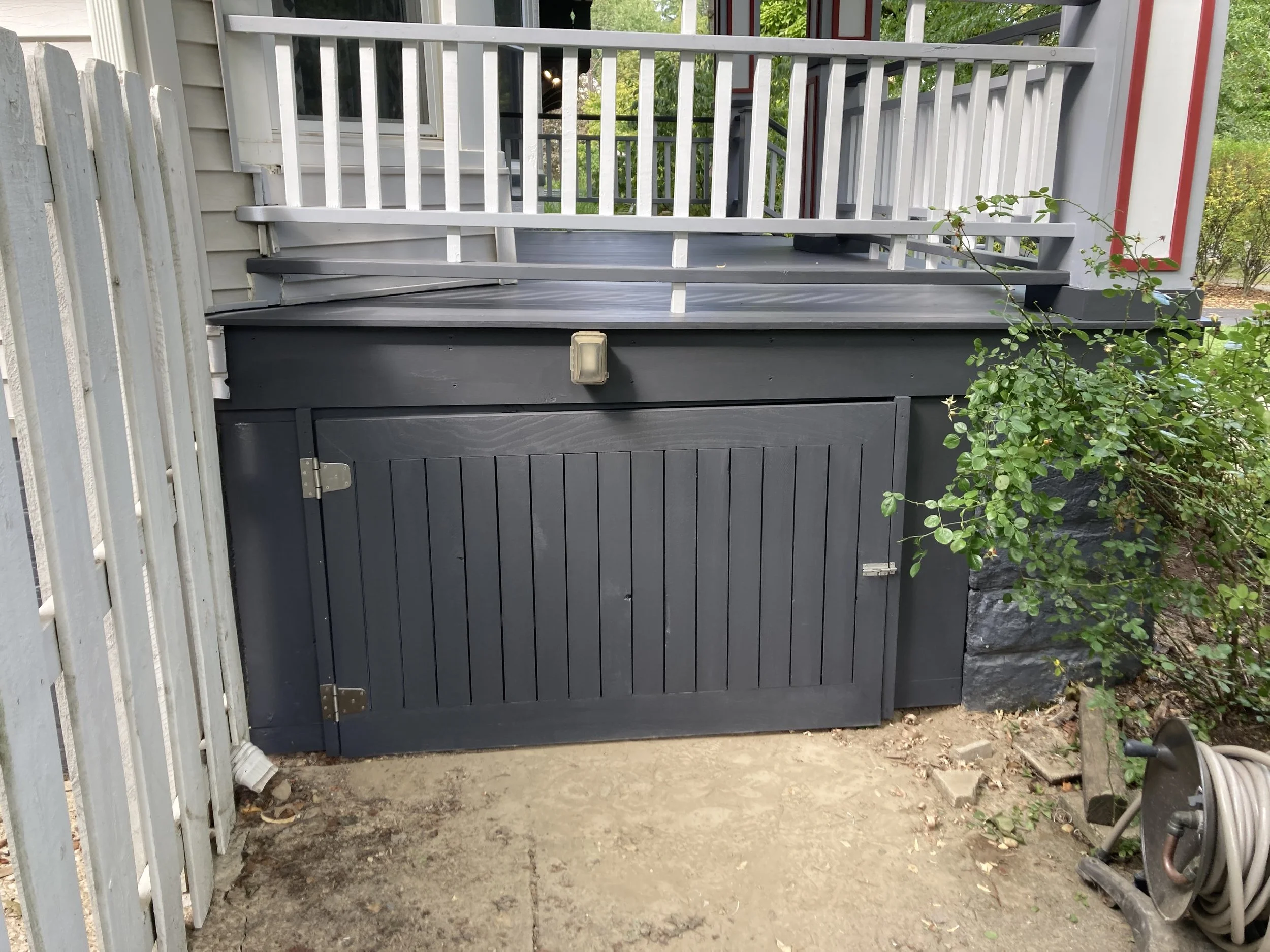 Outdoor storage area with dark wood doors beneath a porch, next to a hose reel and bushes.