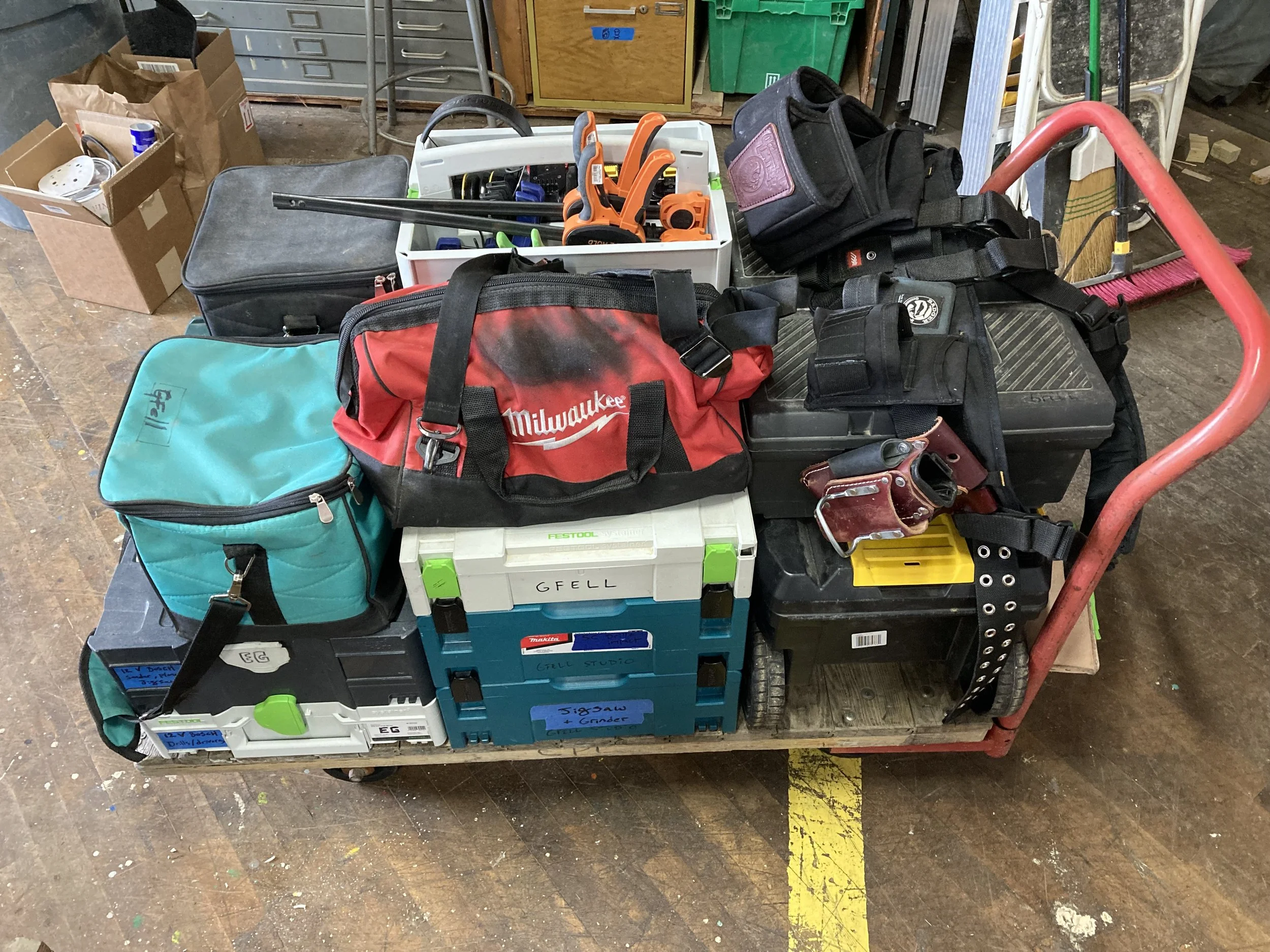 A cart loaded with various tool bags, toolboxes, and equipment, including a Milwaukee bag, a turquoise bag, and several boxes marked "GFELL," indoors on a wooden floor.