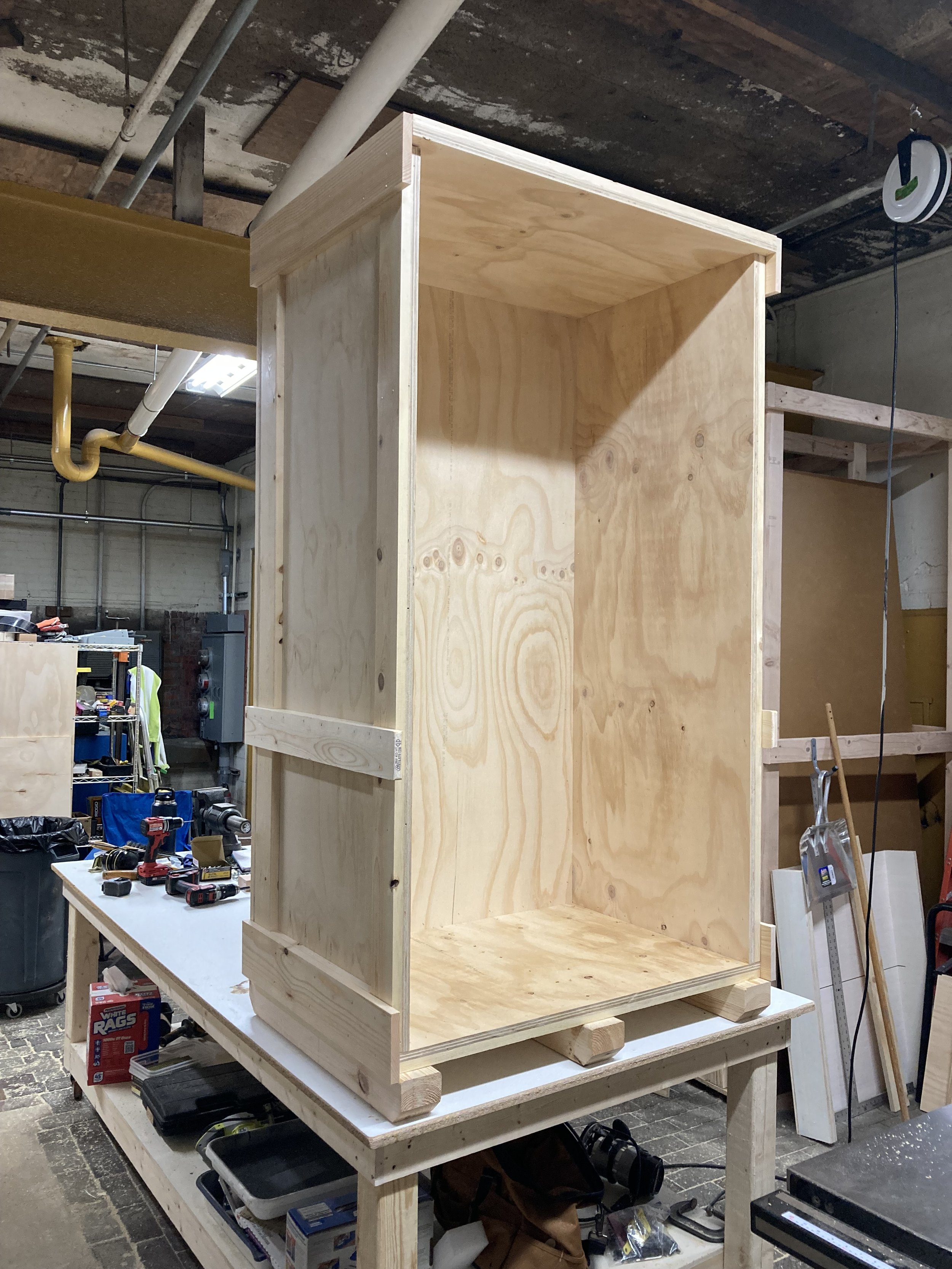 Unfinished wooden cabinet on a workbench in a workshop with tools and materials around.