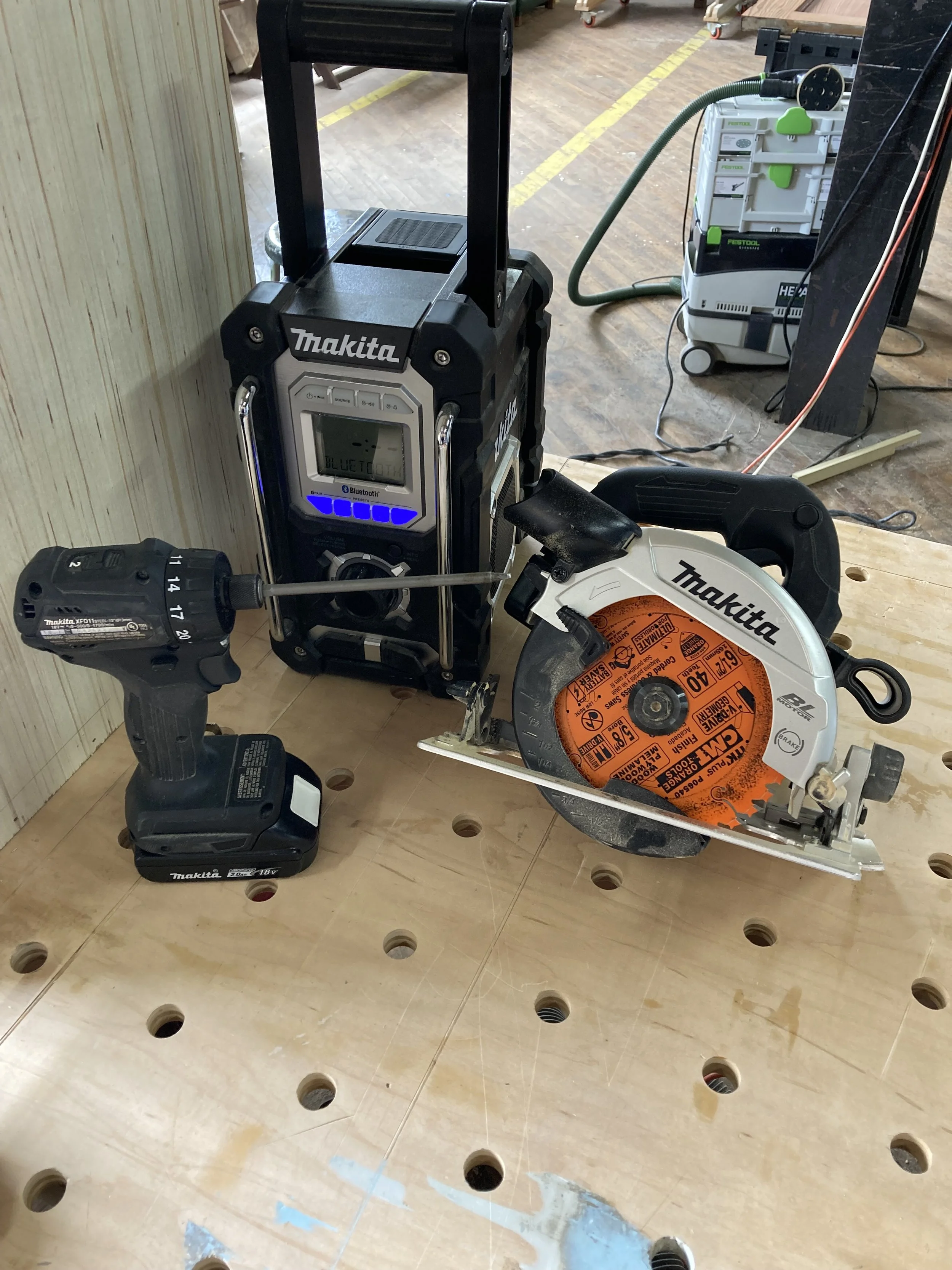 Makita cordless drill, Bluetooth job site radio, and circular saw on a wooden workbench with Festool vacuum in background.