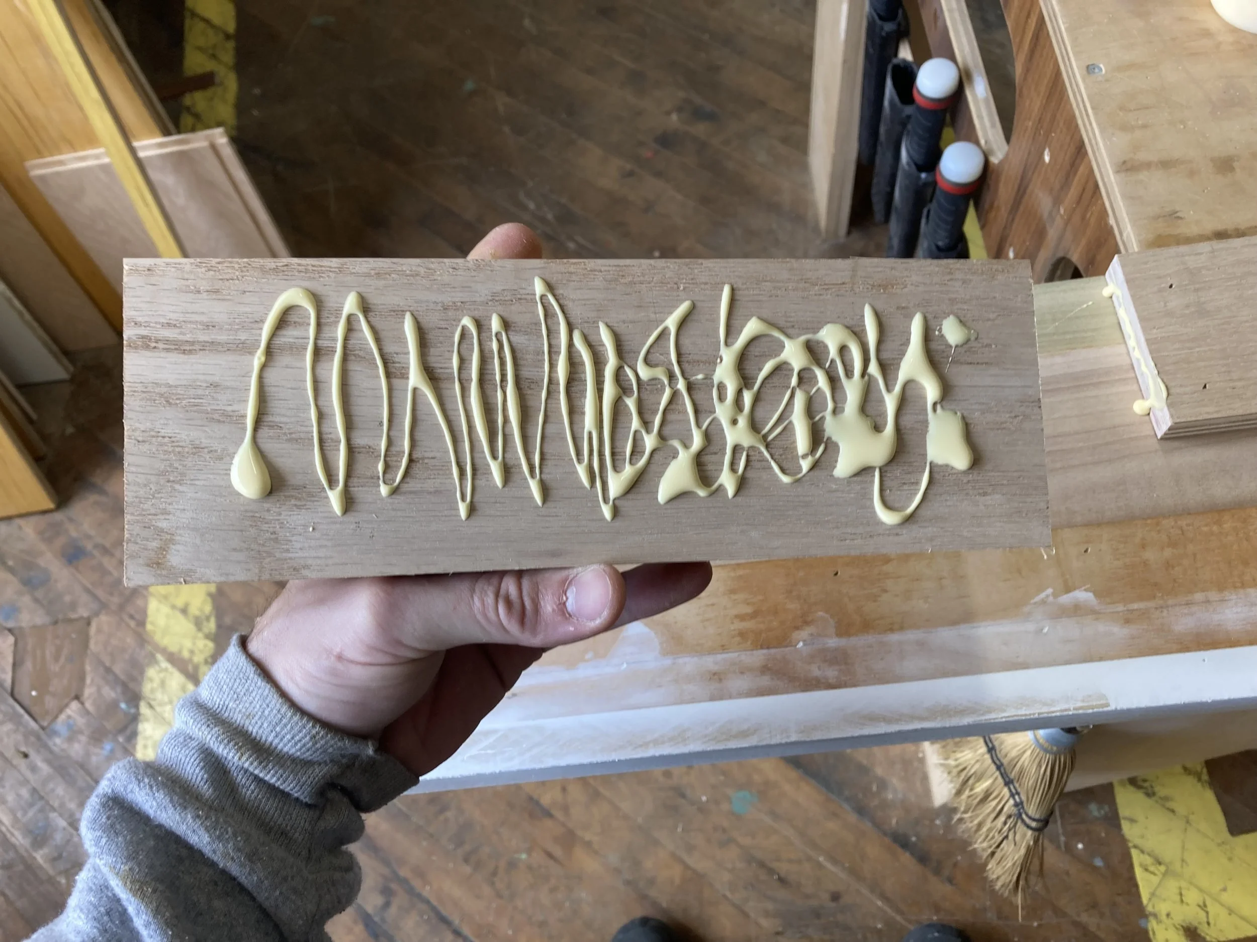 Hand holding a wooden plank with yellow adhesive applied in a wavy pattern, set in a woodworking workshop.