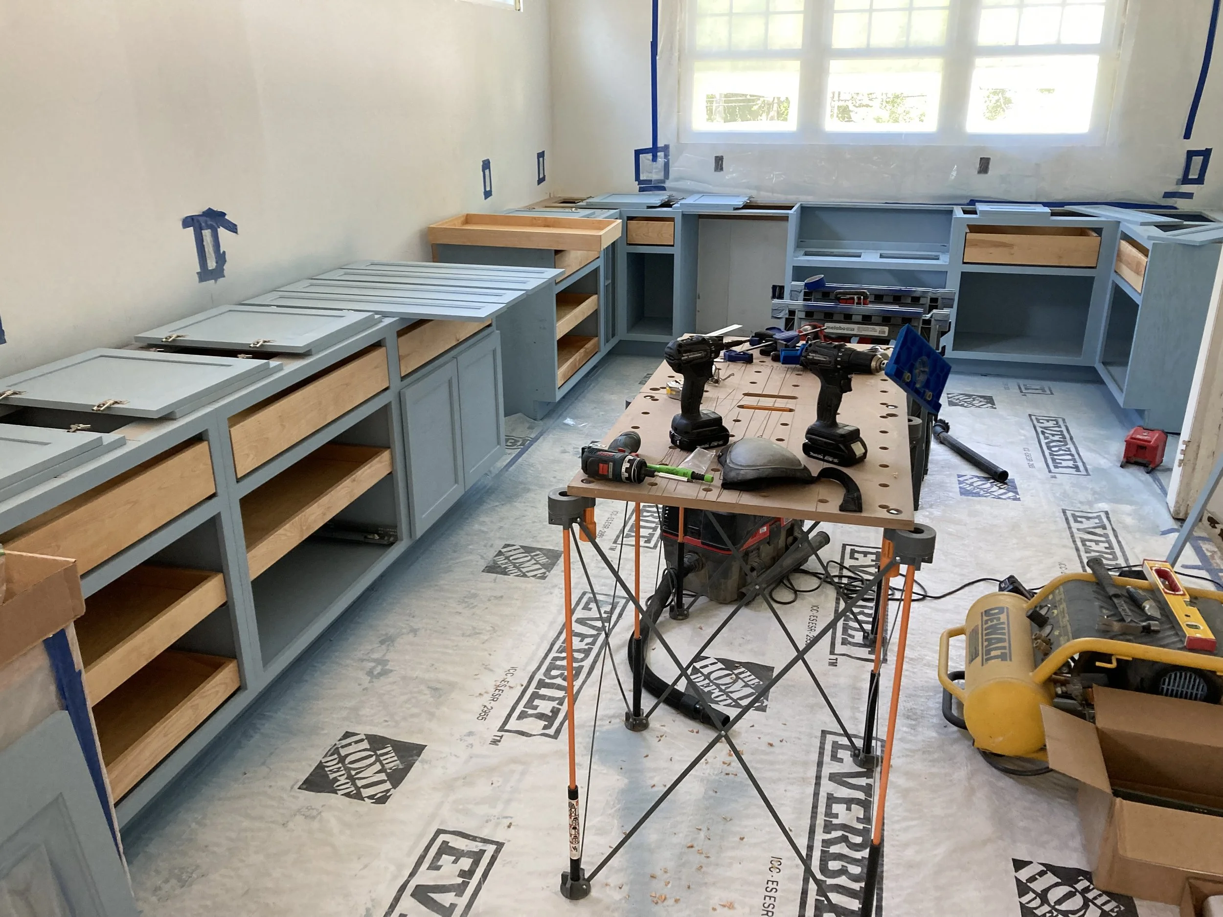 Kitchen under renovation with light blue cabinets being installed, tools and a workbench with power tools present, protective floor covering visible.