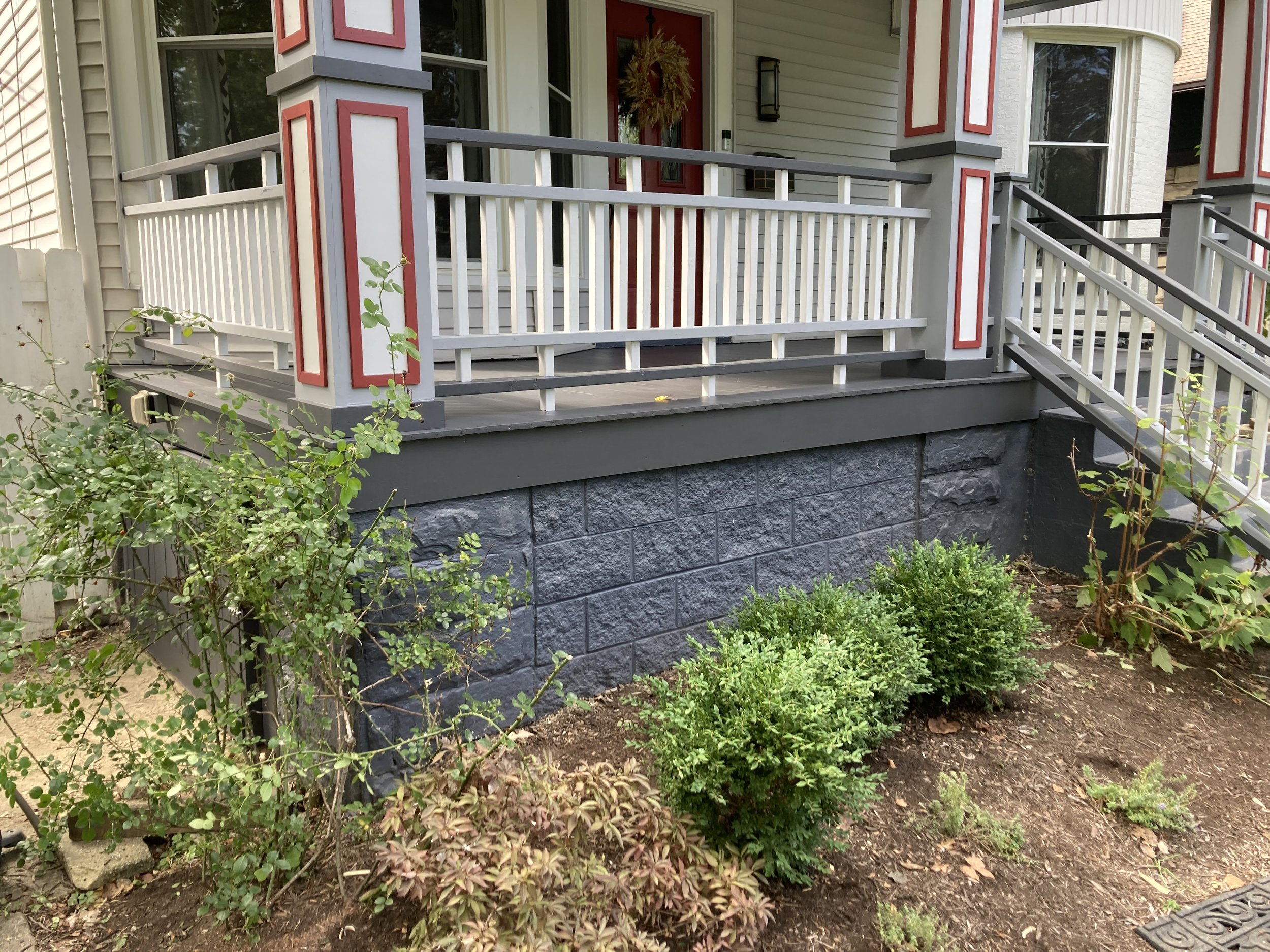 Front porch of a house with white railing, red and white pillars, and gray stone foundation. There are green bushes and plants in a garden bed surrounding the porch.