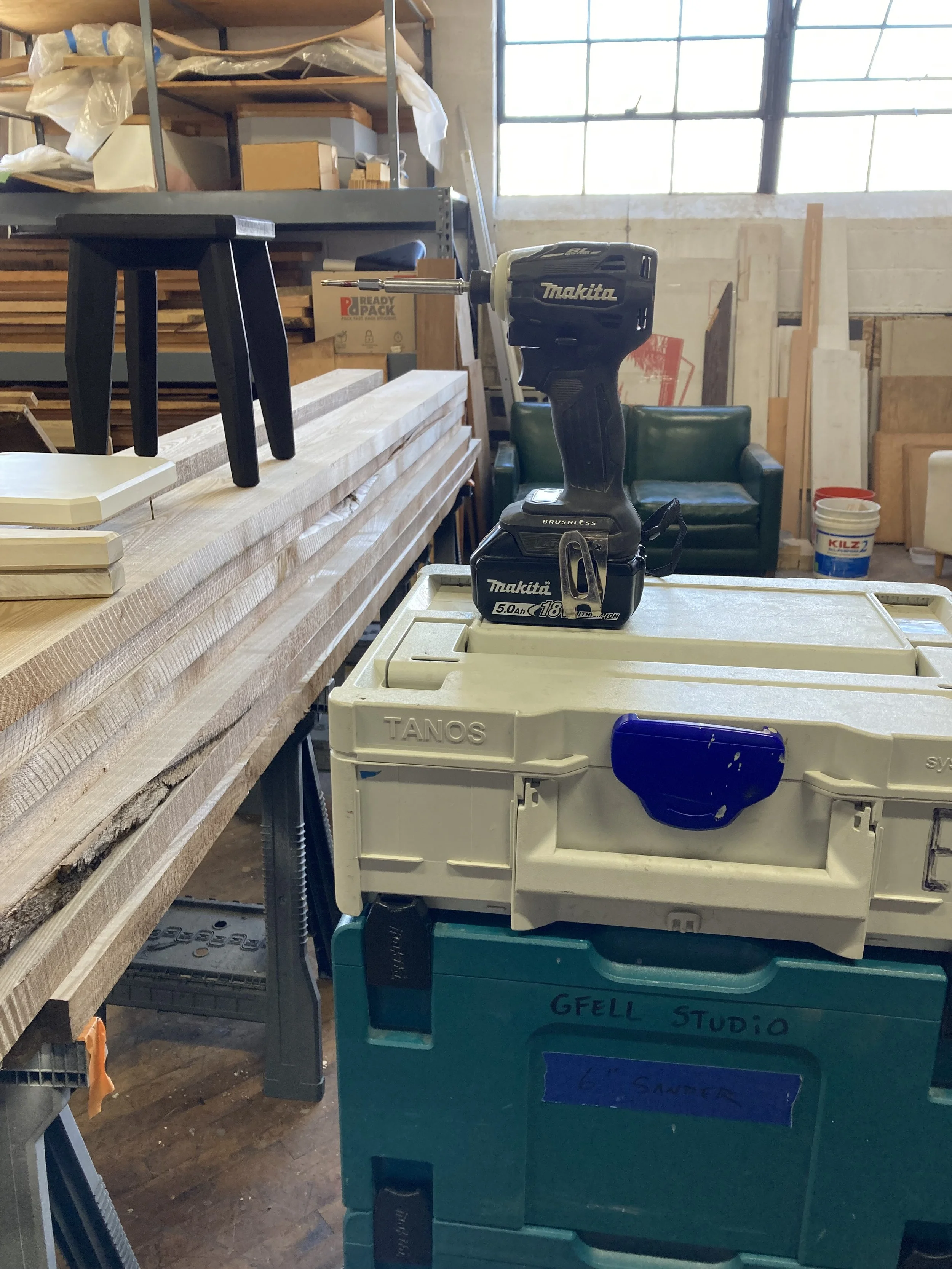 Workshop scene with stacked Makita toolboxes, a Makita drill, wooden boards on a workbench, a black stool, and shelves with materials.