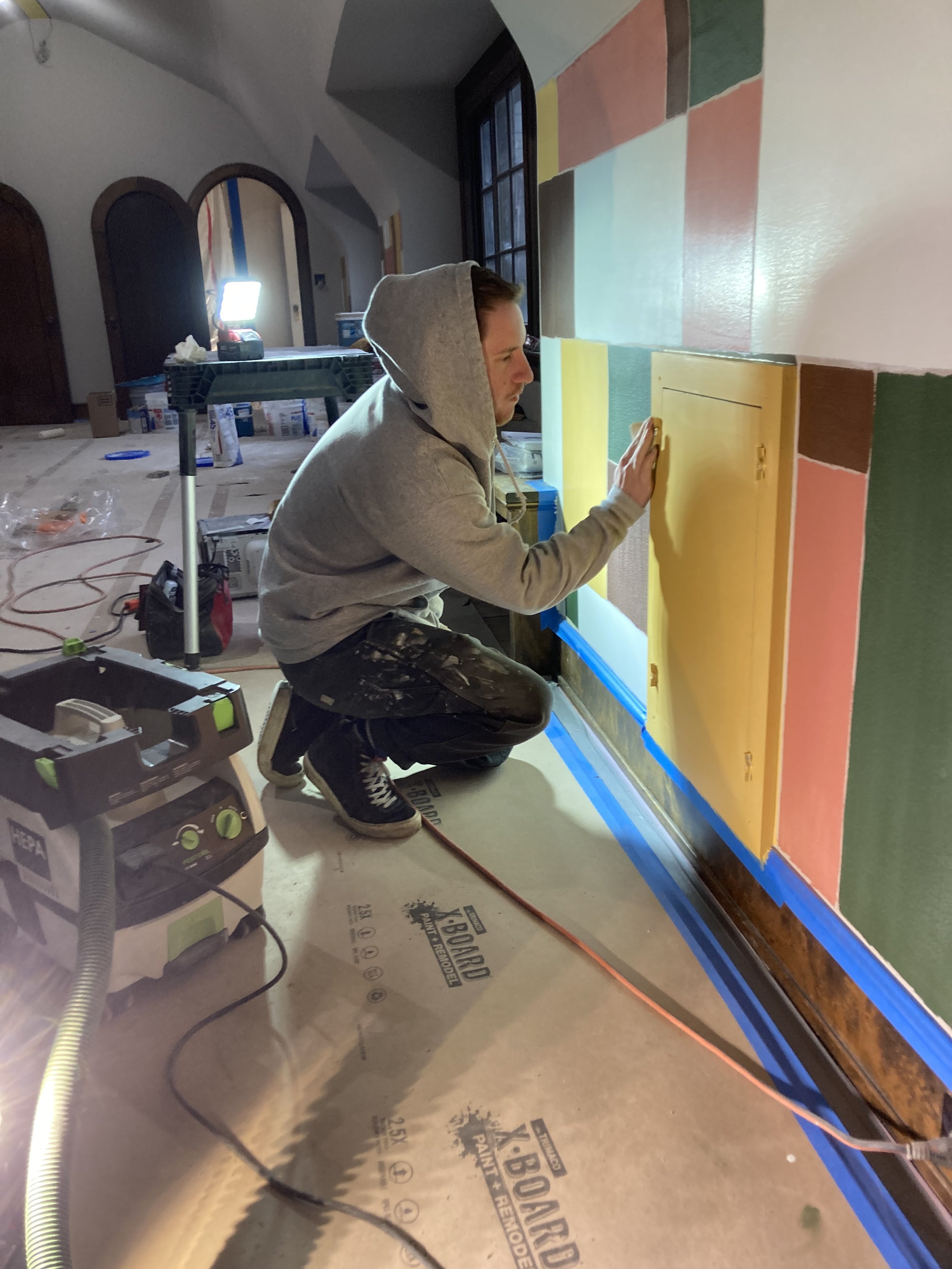 Person in a hoodie kneeling and smoothing a yellow painted panel on a multicolored wall in a room under renovation.