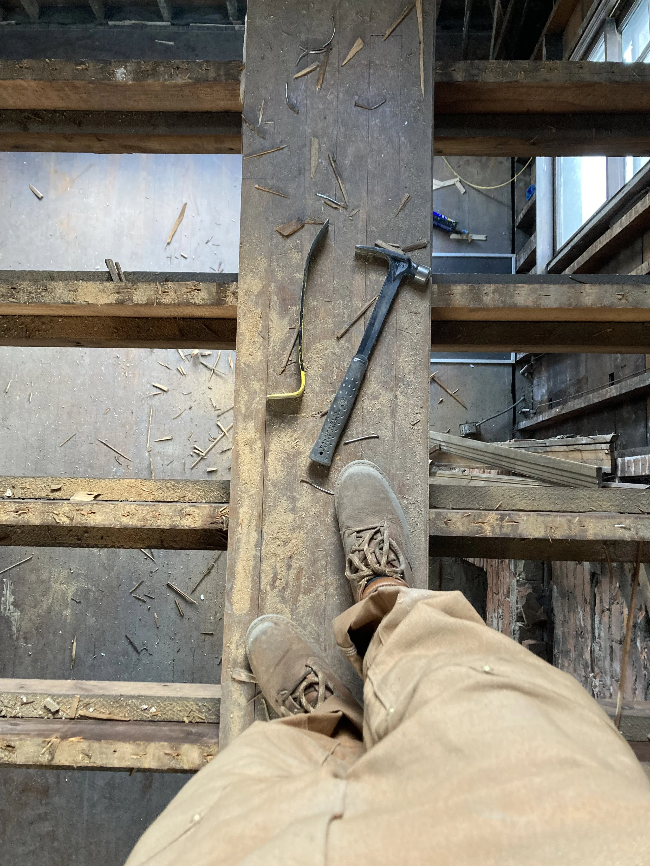 Person standing on a wooden beam with tools, including a hammer and crowbar, visible during a construction or renovation project.