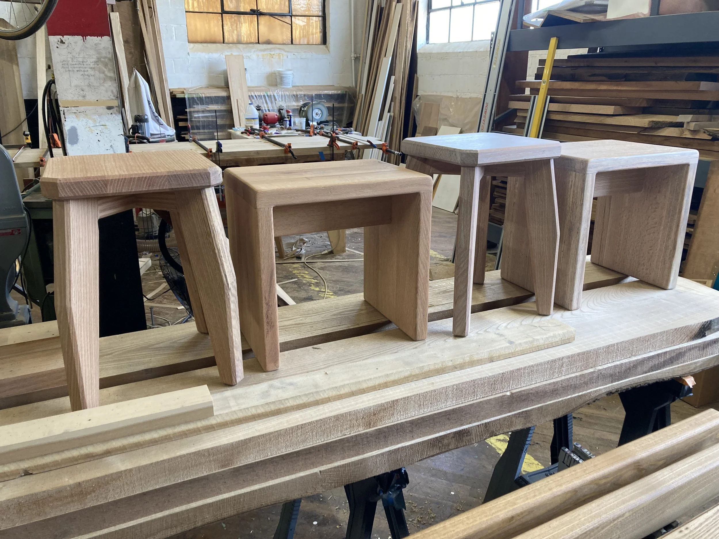 Four wooden stools on a workbench in a woodworking shop.