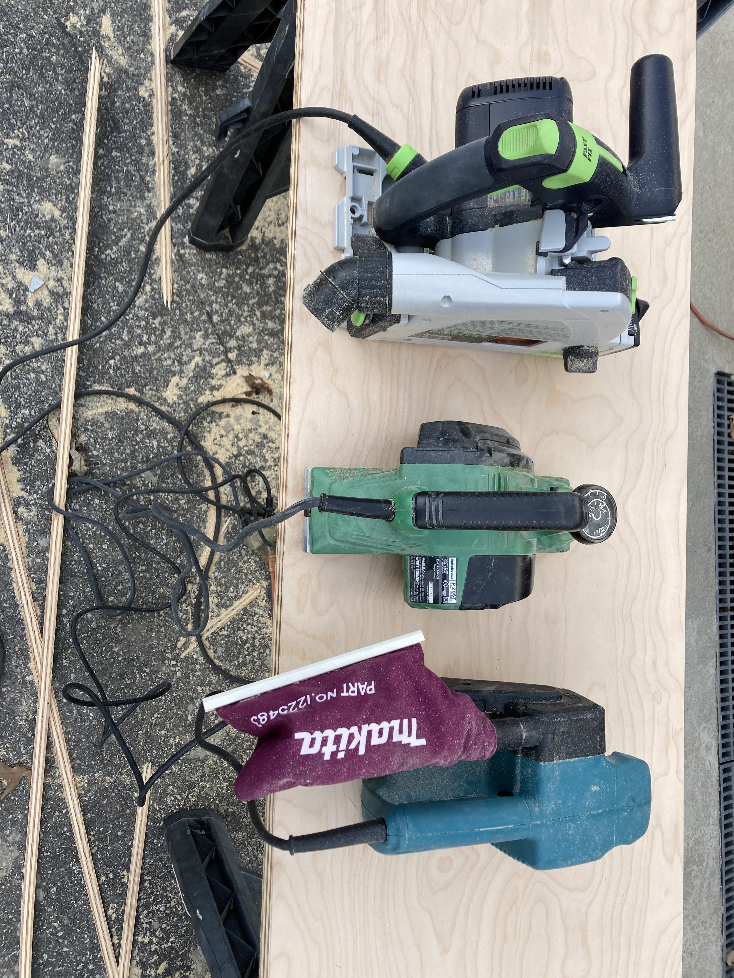 Three power sanders on a wooden surface outdoors with sawdust and wood strips nearby.