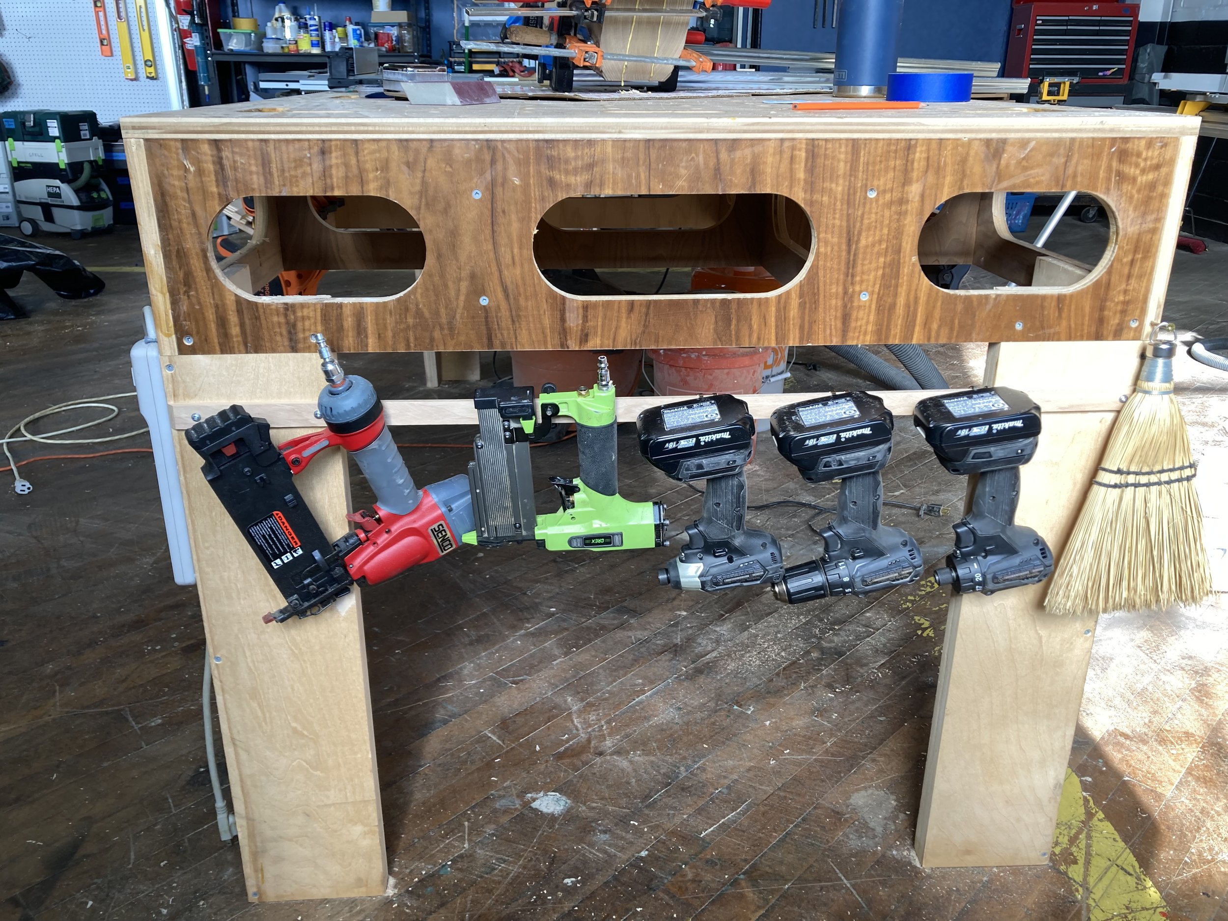 Workbench in a workshop with tools mounted on the side, including drills, nail guns, and a broom.