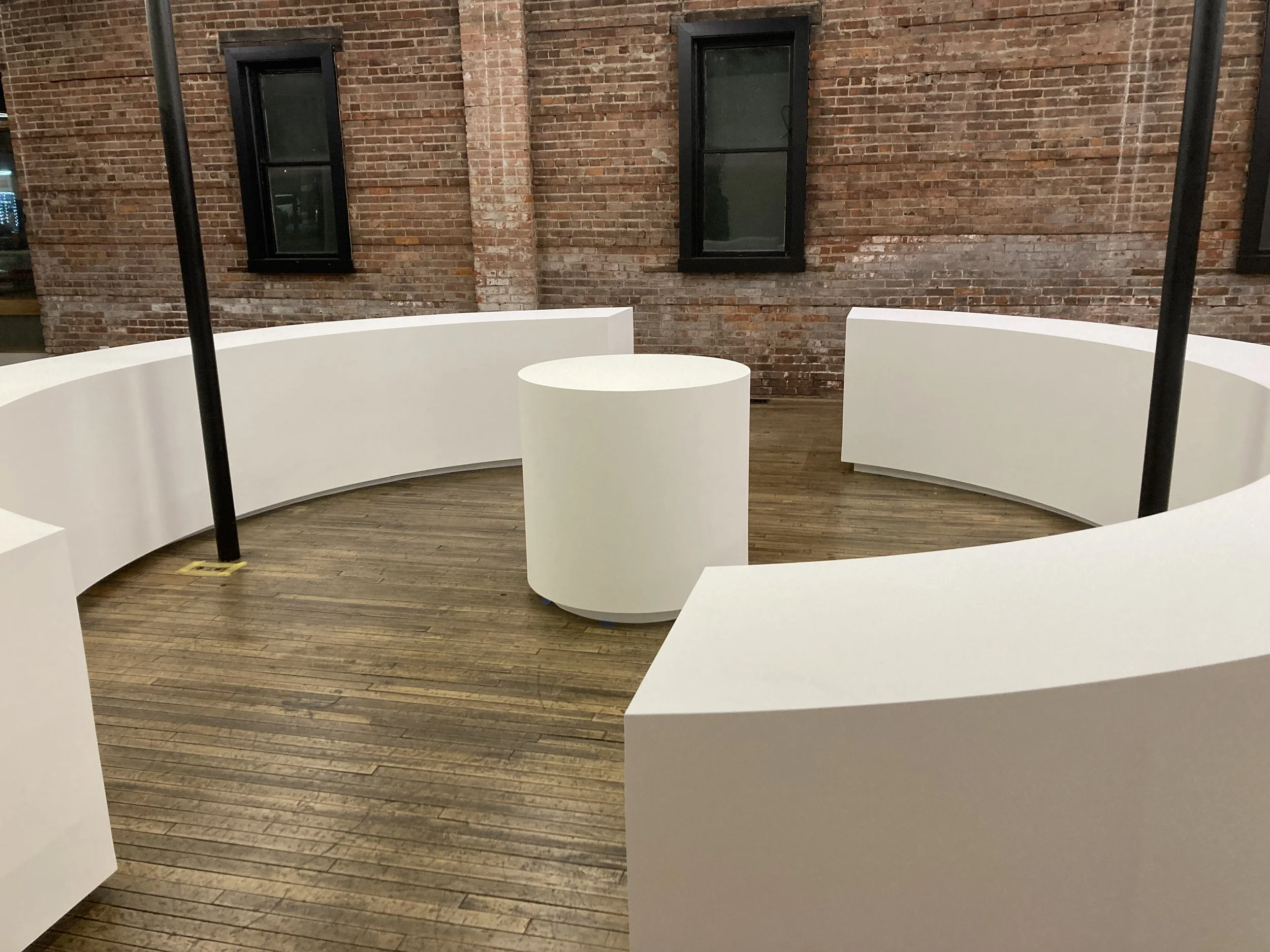 Interior of a room with brick walls and wooden floor, featuring white curved benches arranged in a circle around a cylindrical table.