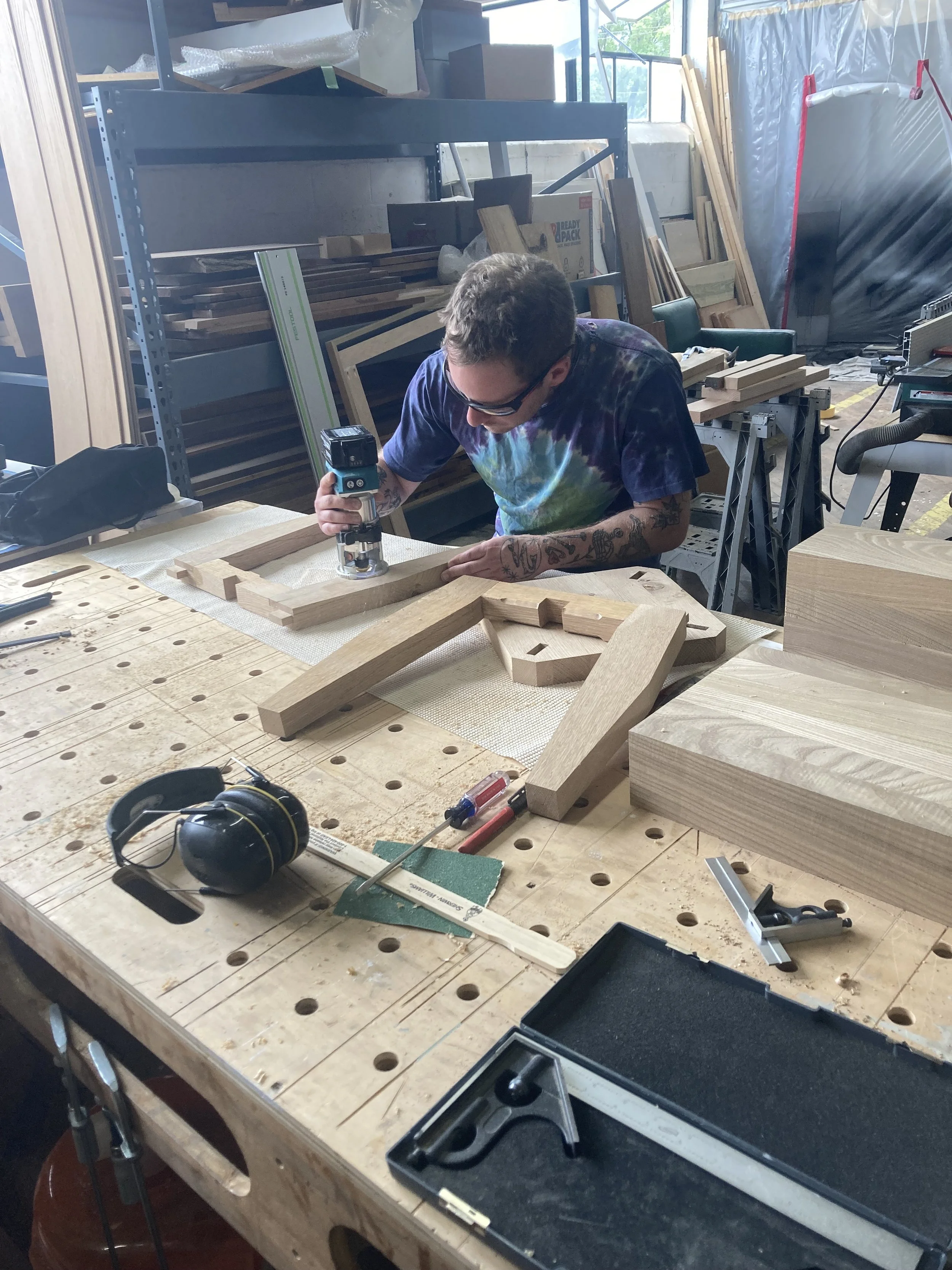 Person using a router on a wooden piece in a workshop, surrounded by tools and woodworking materials.