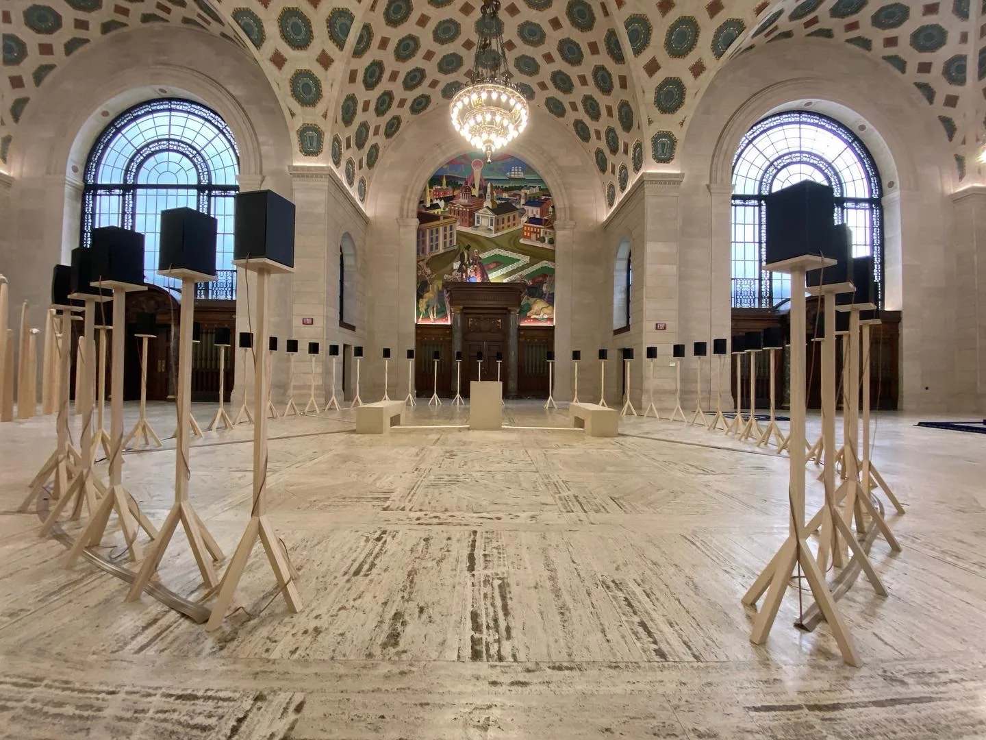 A grand hall with an ornate ceiling featuring decorative patterns and a large chandelier. The room has arched windows with intricate designs and a mural on the far wall. There are multiple stands arranged in a circular pattern on the marble floor.