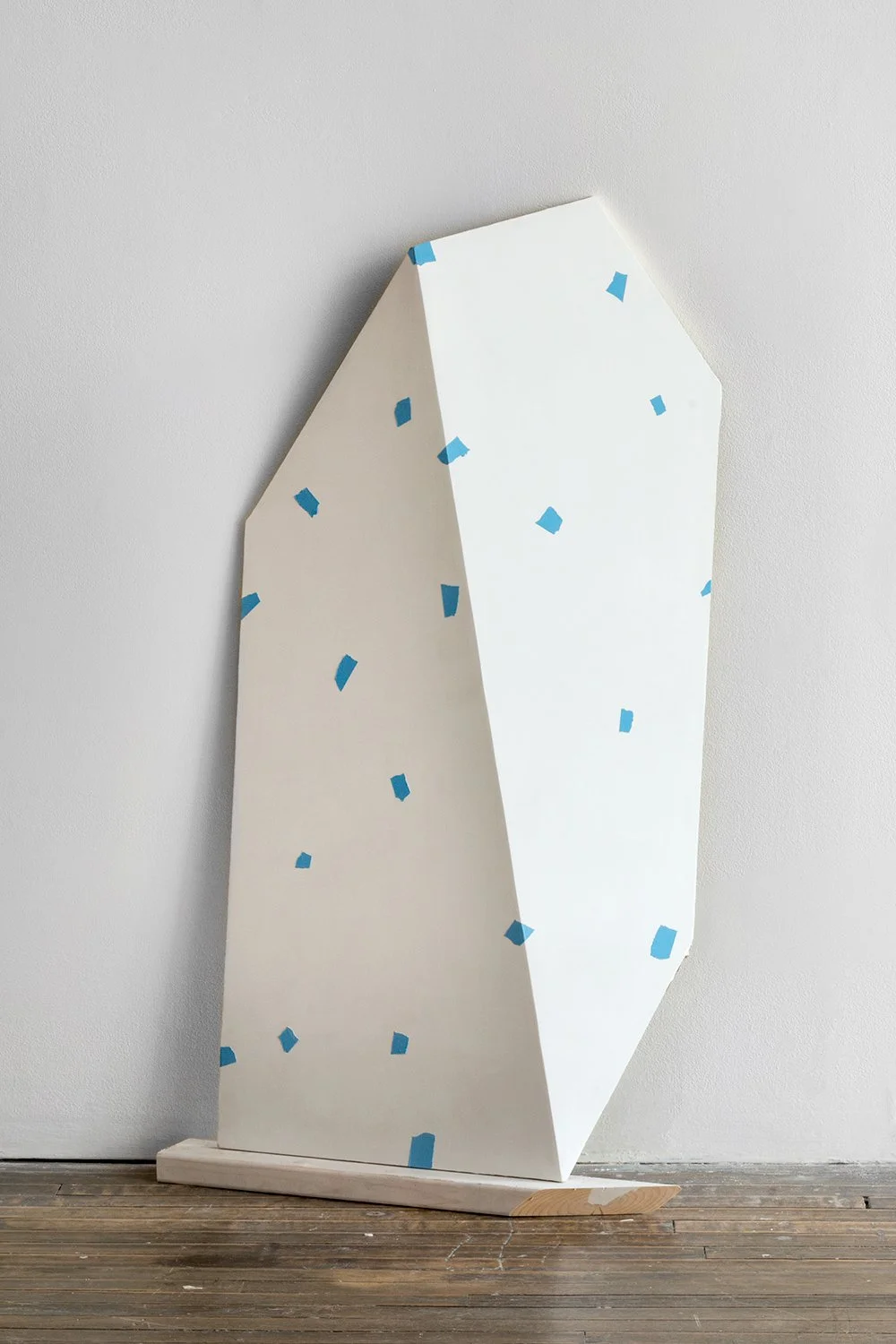 Geometric white sculpture with blue squares on wooden floor against a wall.