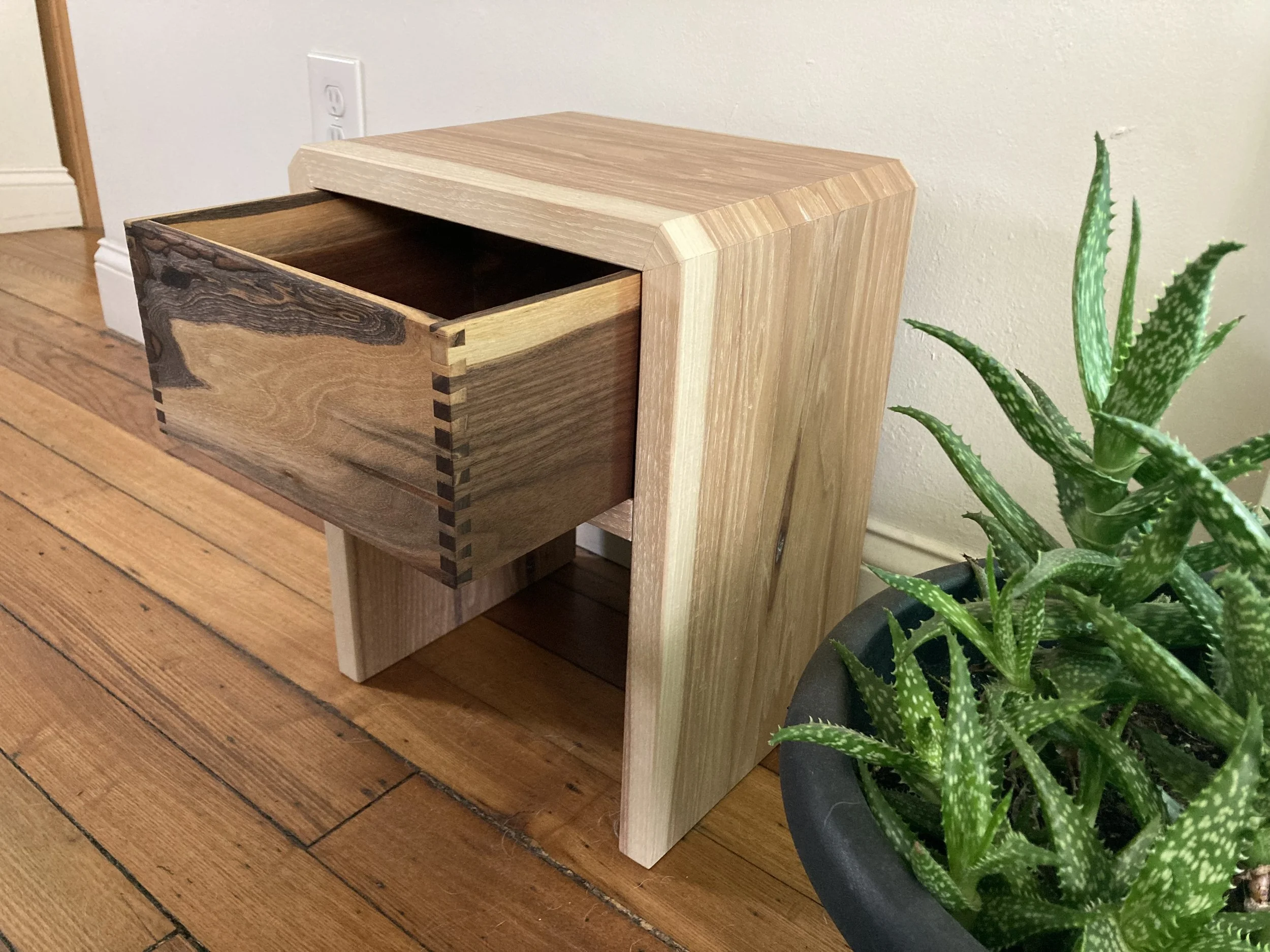 Wooden nightstand with open drawer next to an aloe plant in a black pot on a wood floor.