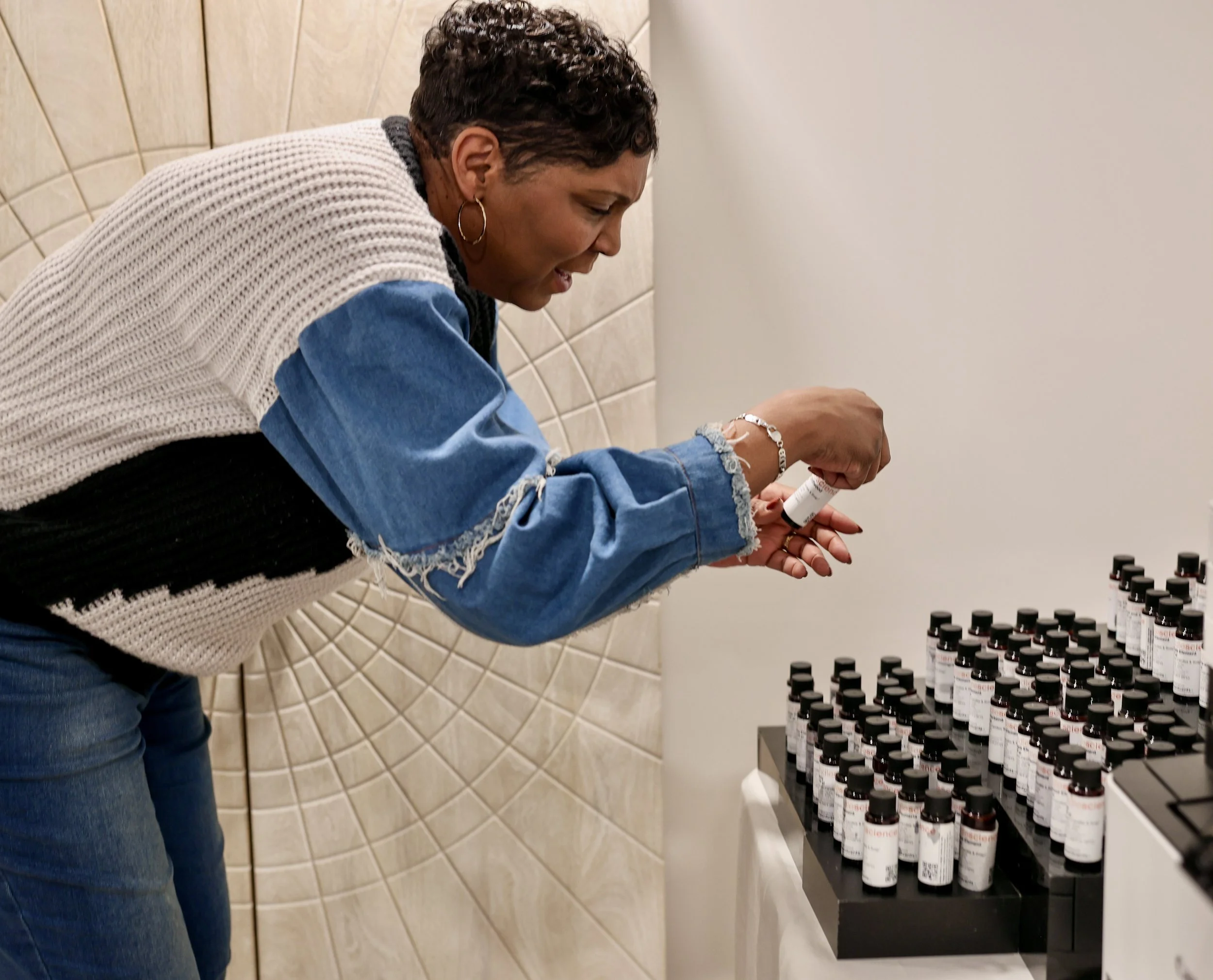 A woman examining small bottles of medication or supplements arranged on a black tray, inside a room with tiled walls.