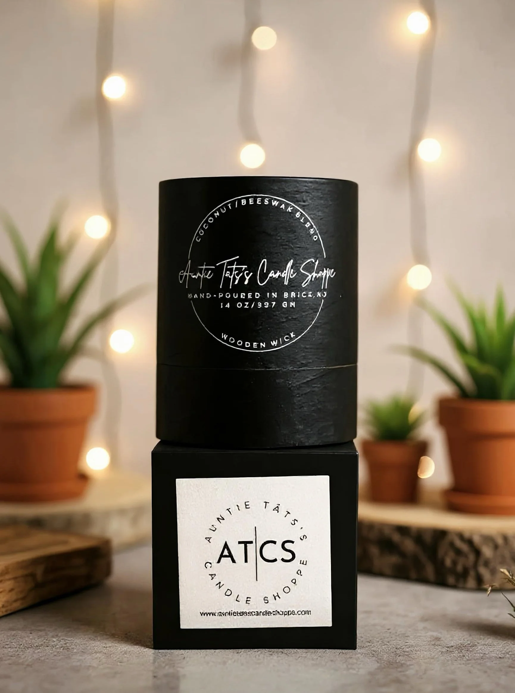 A black cylindrical candle featuring white handwritten text about coconut and beeswax scent, placed on a black box with a white label reading ATCS, and surrounded by decorative plants and string lights in the background.