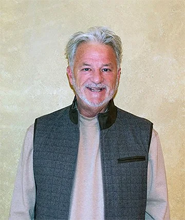 A smiling middle-aged man with gray hair and beard, wearing a beige shirt and a dark gray vest, standing against a plain light-colored wall.