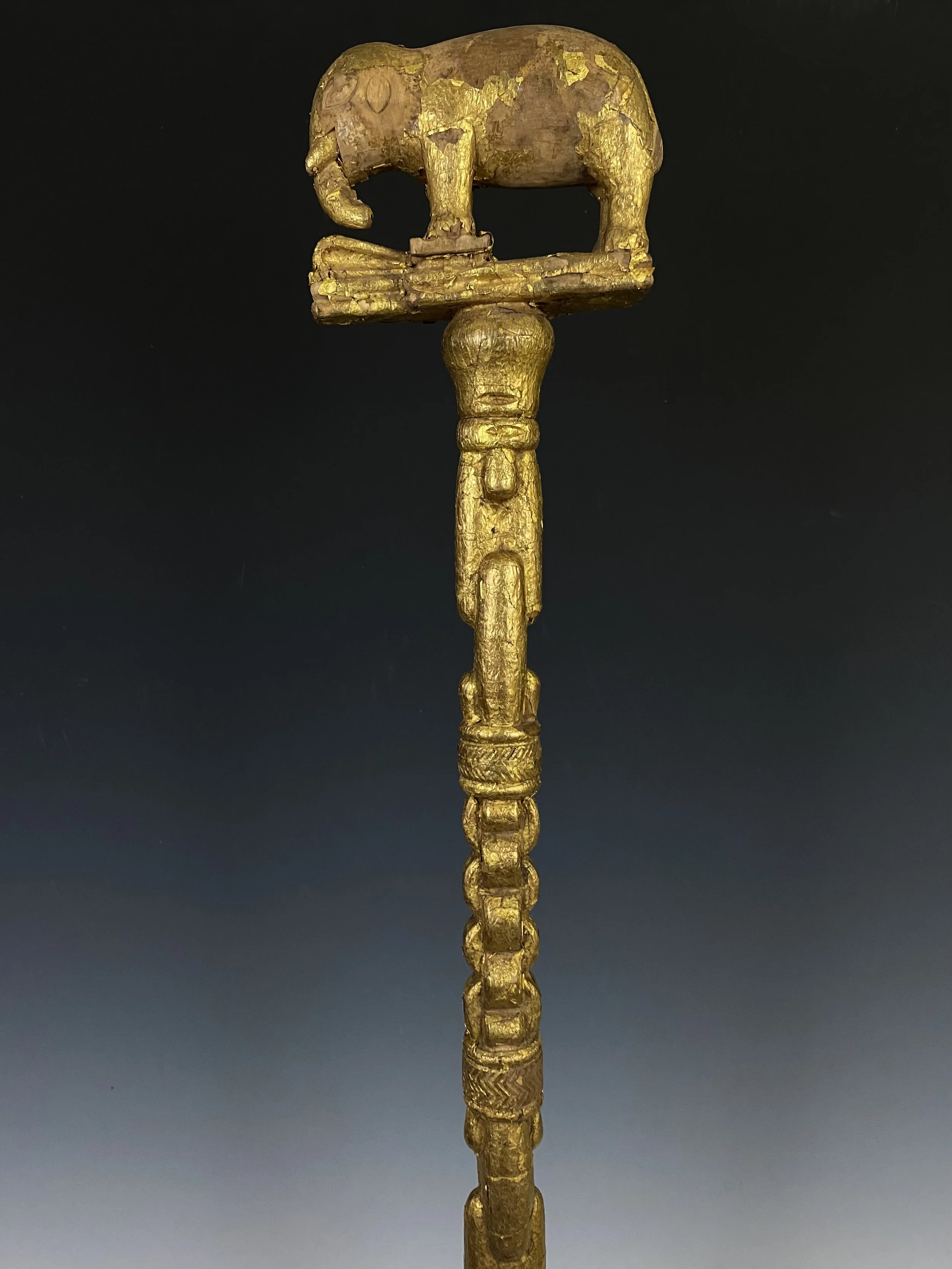 An Asante linguist/ counselor’s staff topped with a finial of an elephant stepping on a trap. Wood, covered with gold foil, Ghana. 43.5” H. The staff is too short to be complete and is most likely missing the middle section as evident in the miss mat