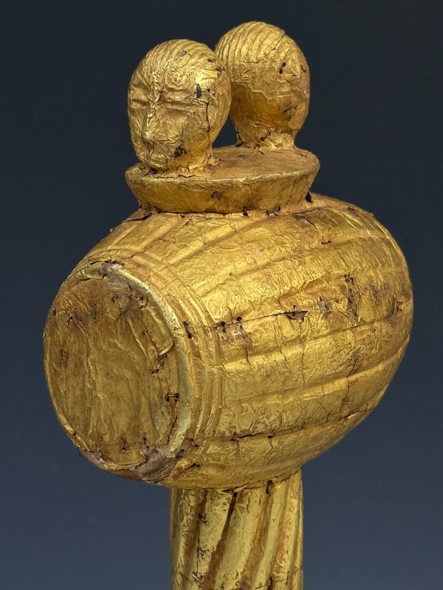 An Akan sculptural sword hilt carved of wood and covered with gold foil and gold staples. 23 cm. Ghana. The image consist of two heads on top of two gunpowder keg barrels- a reference to the power of the state and chief.