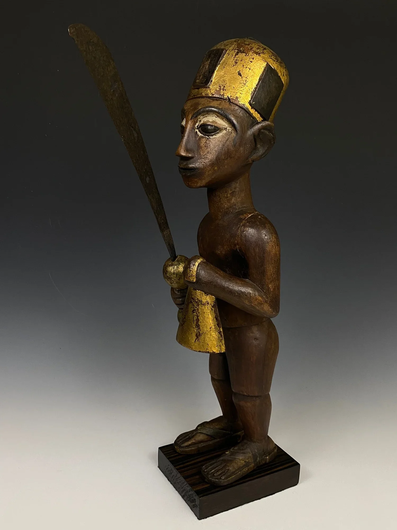 Asante shrine figure of a ruler. Ghana. Wood, pigments, kaolin (white clay), gold leaf, steel. 21'' x 6.5'' x 6''. The ruler is depicted holding a sword (akrafena) with cut adinkra symbols and a gong. He is wearing a traditional abotire with protecti