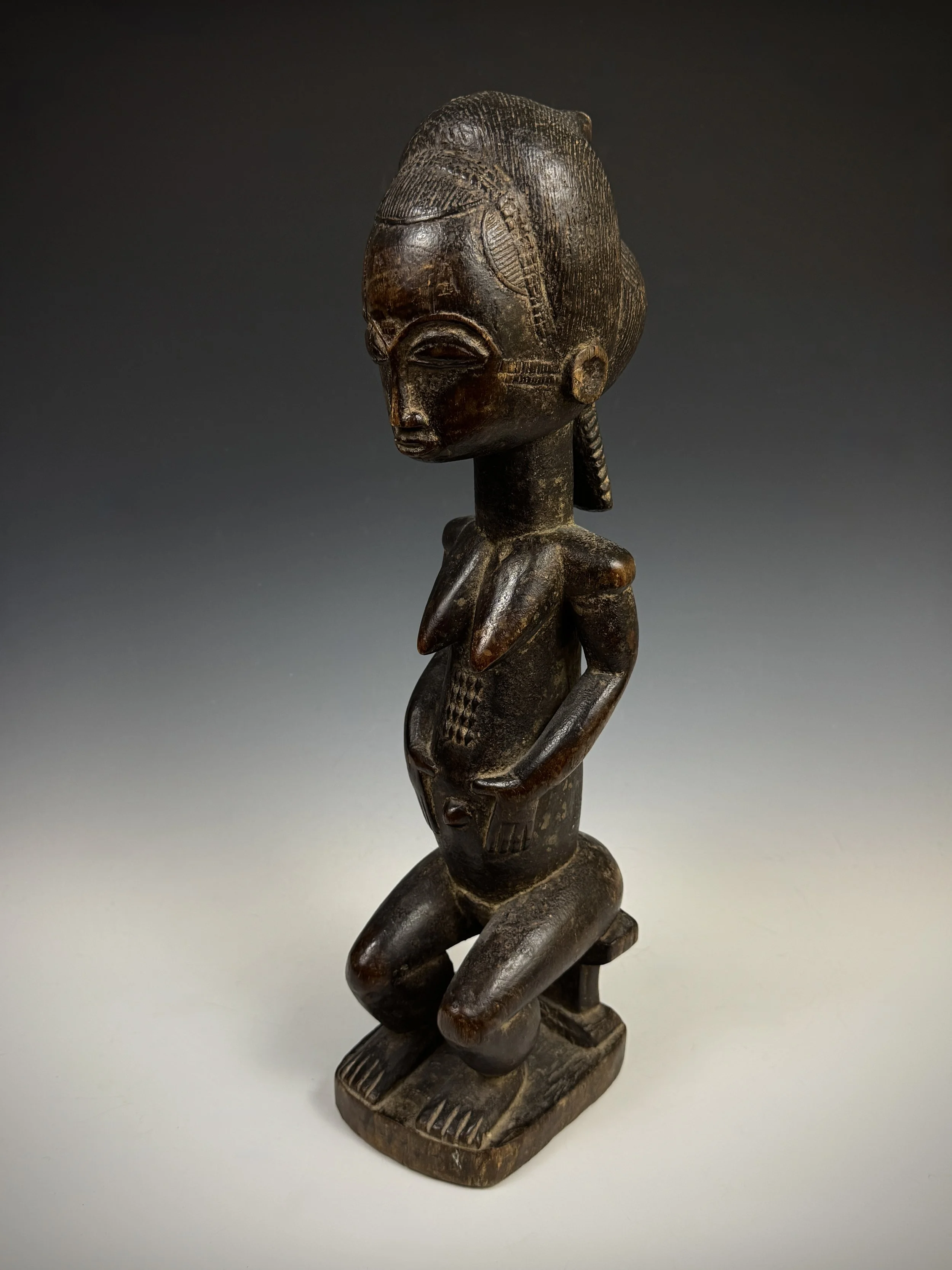  A Baule carved wood female figure (blolo ba) shown seated on a stool,  Côte d'Ivoire.  Size: 18 1/4" L, Weight: 3 Ib. 5.8 oz. “Spirit mate” figures are carved after consultation with a diviner and lives with the owner.  Every Baule had a spirit mate