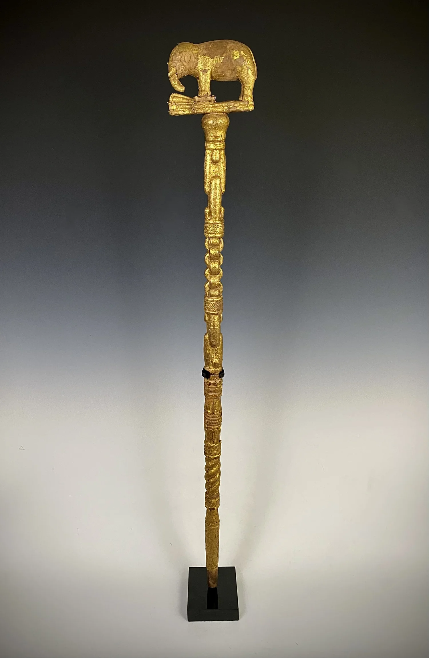 An Asante linguist/ counselor’s staff topped with a finial of an elephant stepping on a trap. Wood, covered with gold foil, Ghana. 43.5” H. The staff is too short to be complete and is most likely missing the middle section as evident in the miss mat