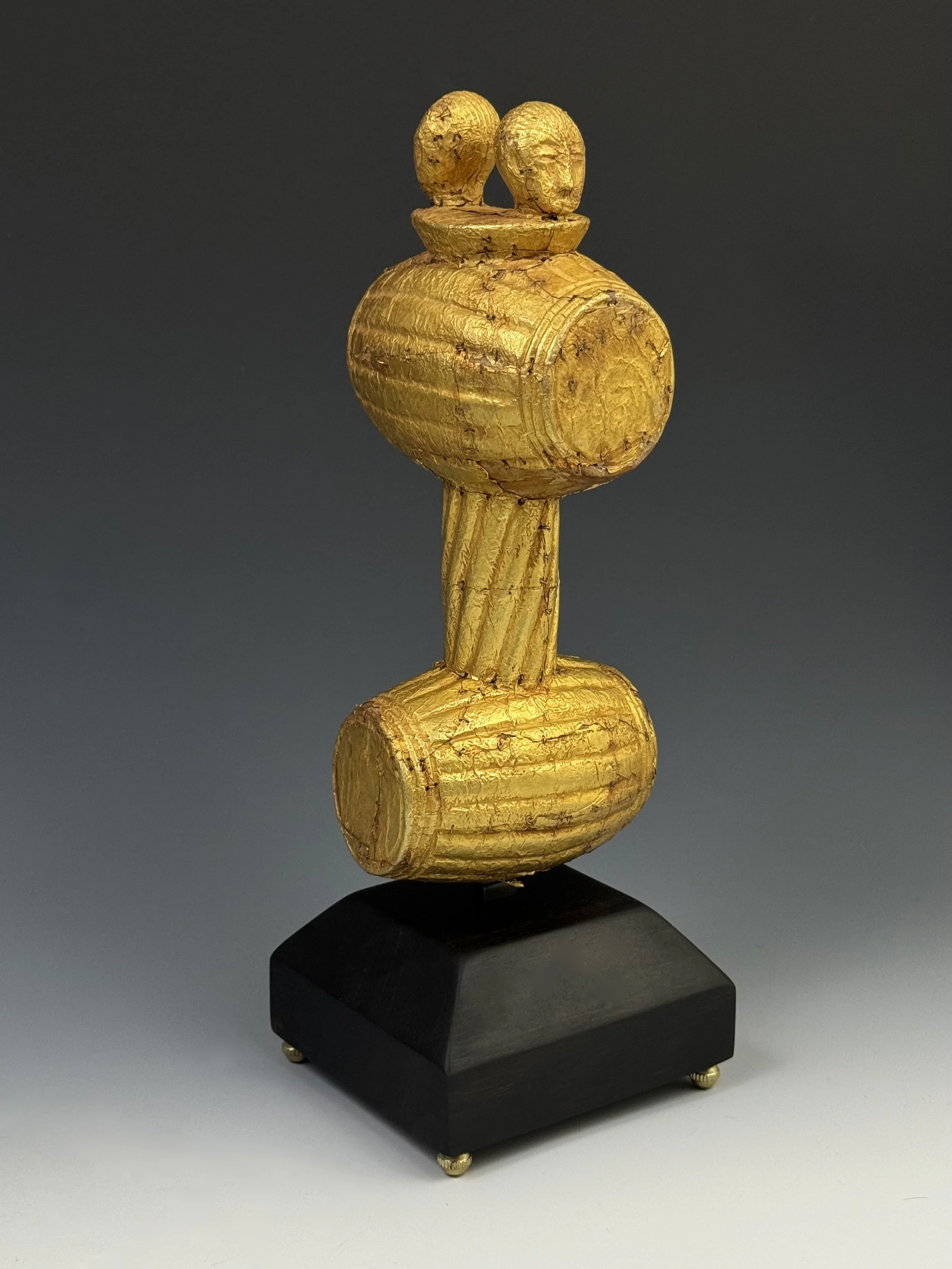 An Akan sculptural sword hilt carved of wood and covered with gold foil and gold staples. 23 cm. Ghana. The image consist of two heads on top of two gunpowder keg barrels- a reference to the power of the state and chief.