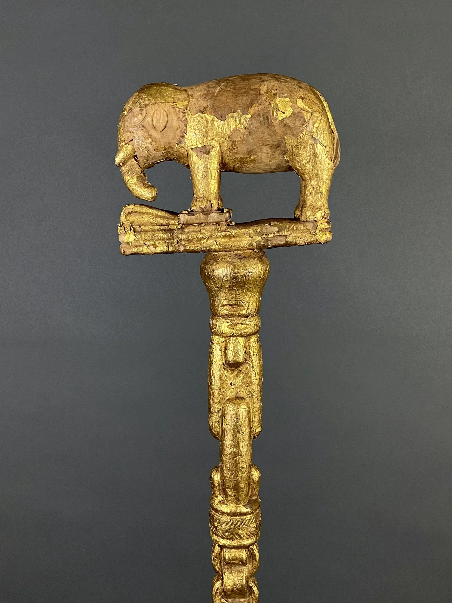 An Asante linguist/ counselor’s staff topped with a finial of an elephant stepping on a trap. Wood, covered with gold foil, Ghana. 43.5” H. The staff is too short to be complete and is most likely missing the middle section as evident in the miss mat
