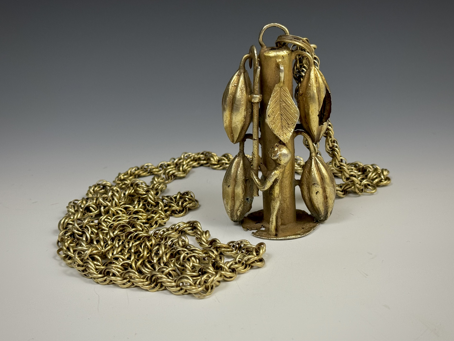 An Akan silver/gold alloy pendant with a very long multi link chain featuring a large depiction of a man picking cacao pods from a tree (321 grams). Size 74" L, pendant 4" H. Silver alloyed with gold is often referred to as "green gold". When it natu