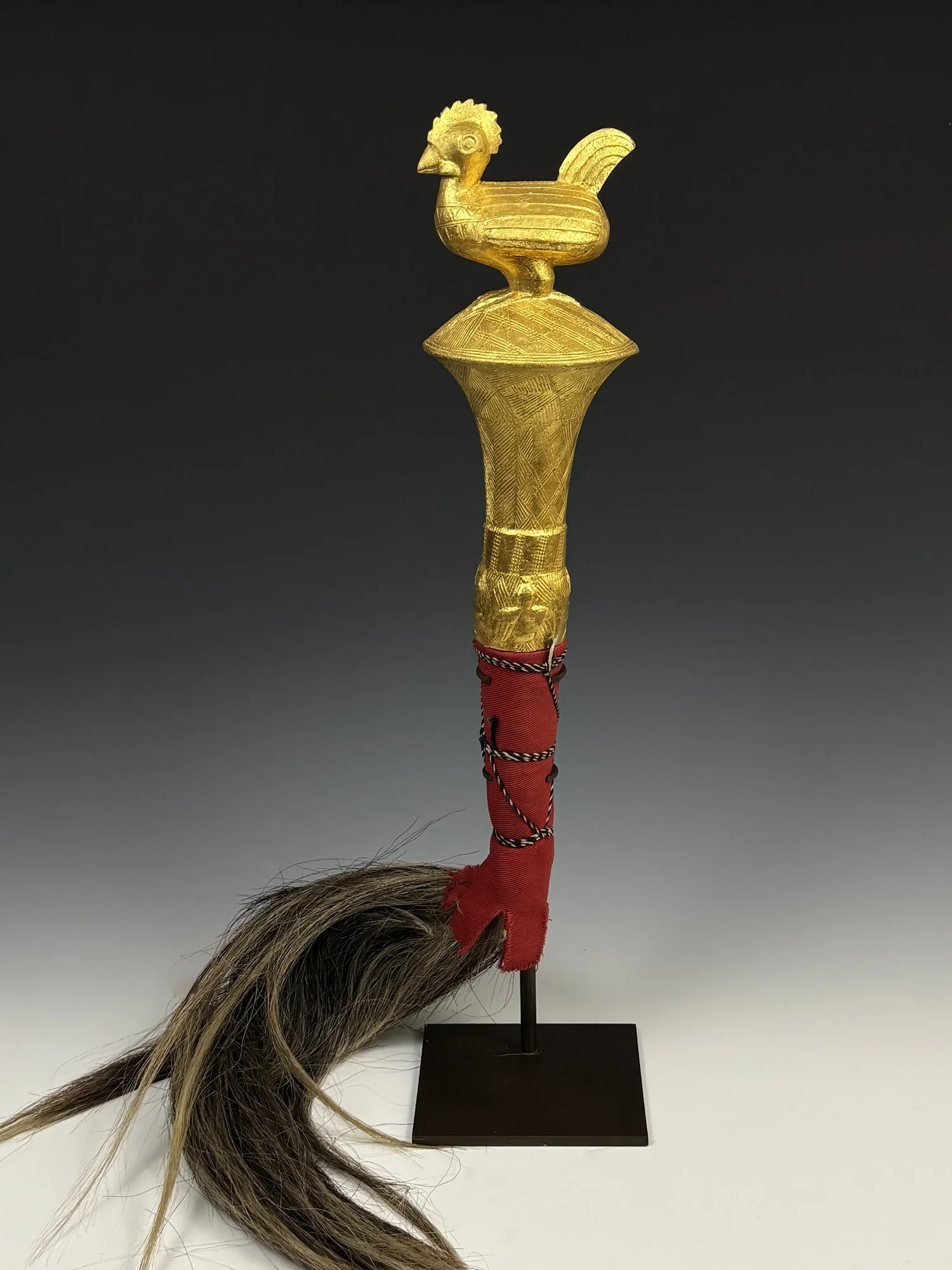 Baule fly whisk. Handle: 40cm H. Wood covered with gold foil, horse hair, textile, Côte d’Ivoire. The finial image is of a standing rooster. “Akokg ntakdra na emd akoko ye kese”, —It is the feathers on a fowl that make it big. (This saying has been h