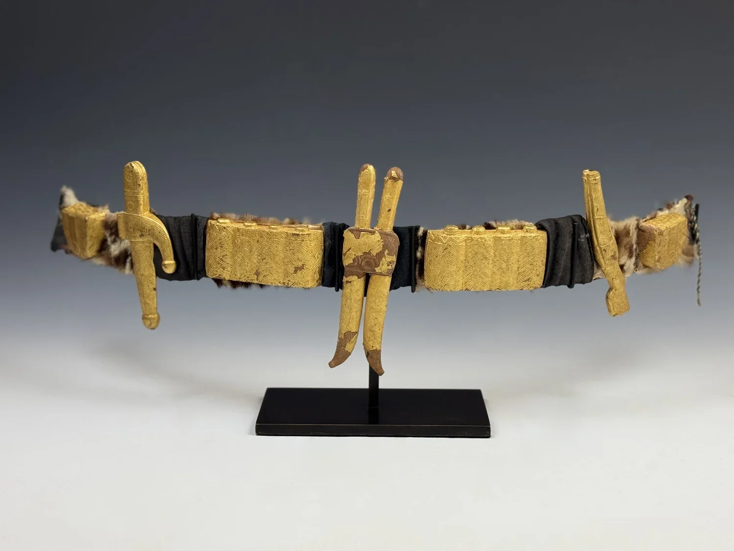 An Akan belt of a notable, Côte d'Ivoire. Wood, covered with gold foil, fur, textile. L 72cm. It features the distinctive double-knife that illustrates a Baule proverb, ““The blade of a knife cannot carve its own handle,” demonstrating the need for c