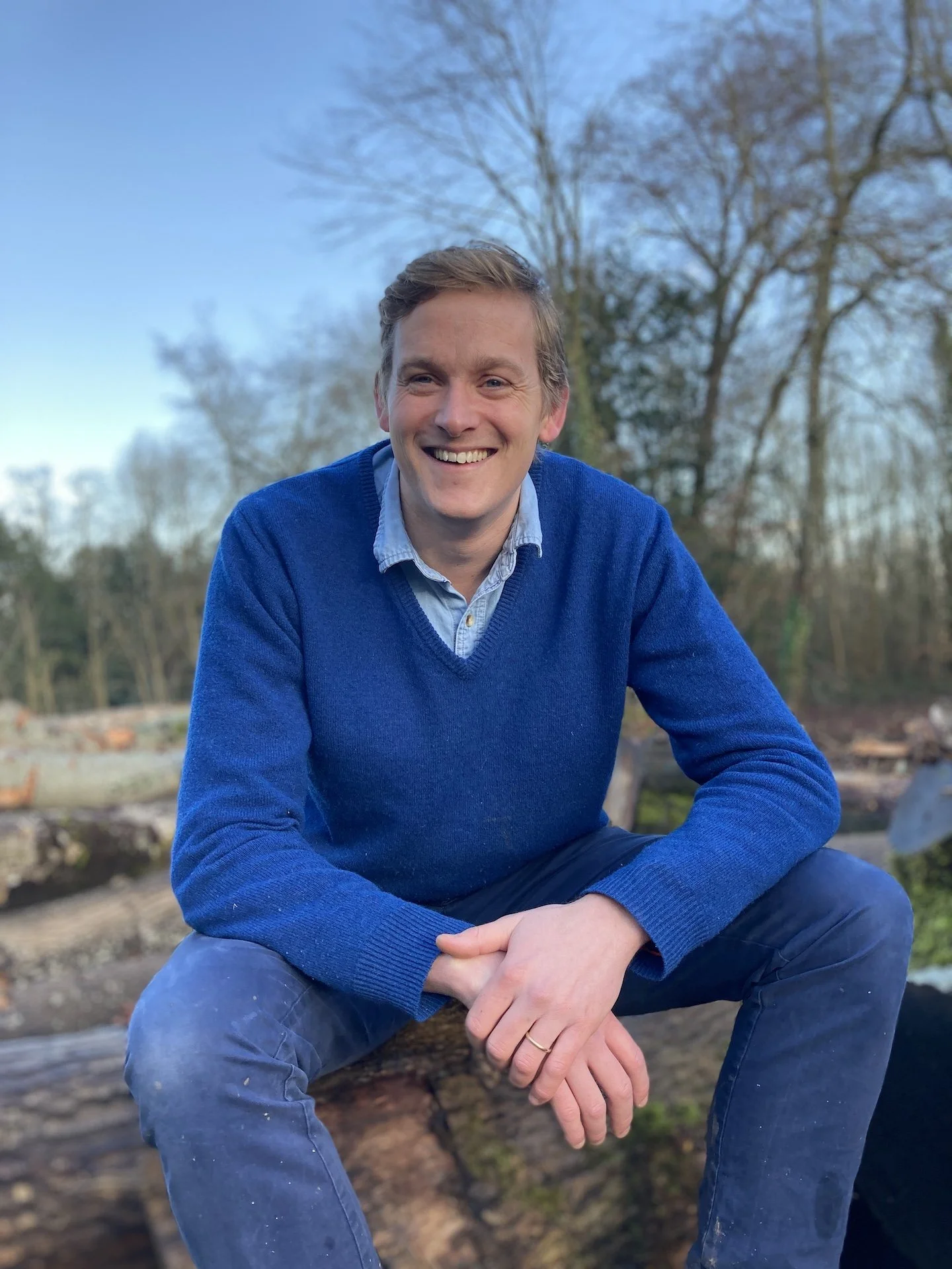 A man sitting on a woodworking workbench in a workshop, surrounded by tools and equipment, wearing a work apron and casual clothing.