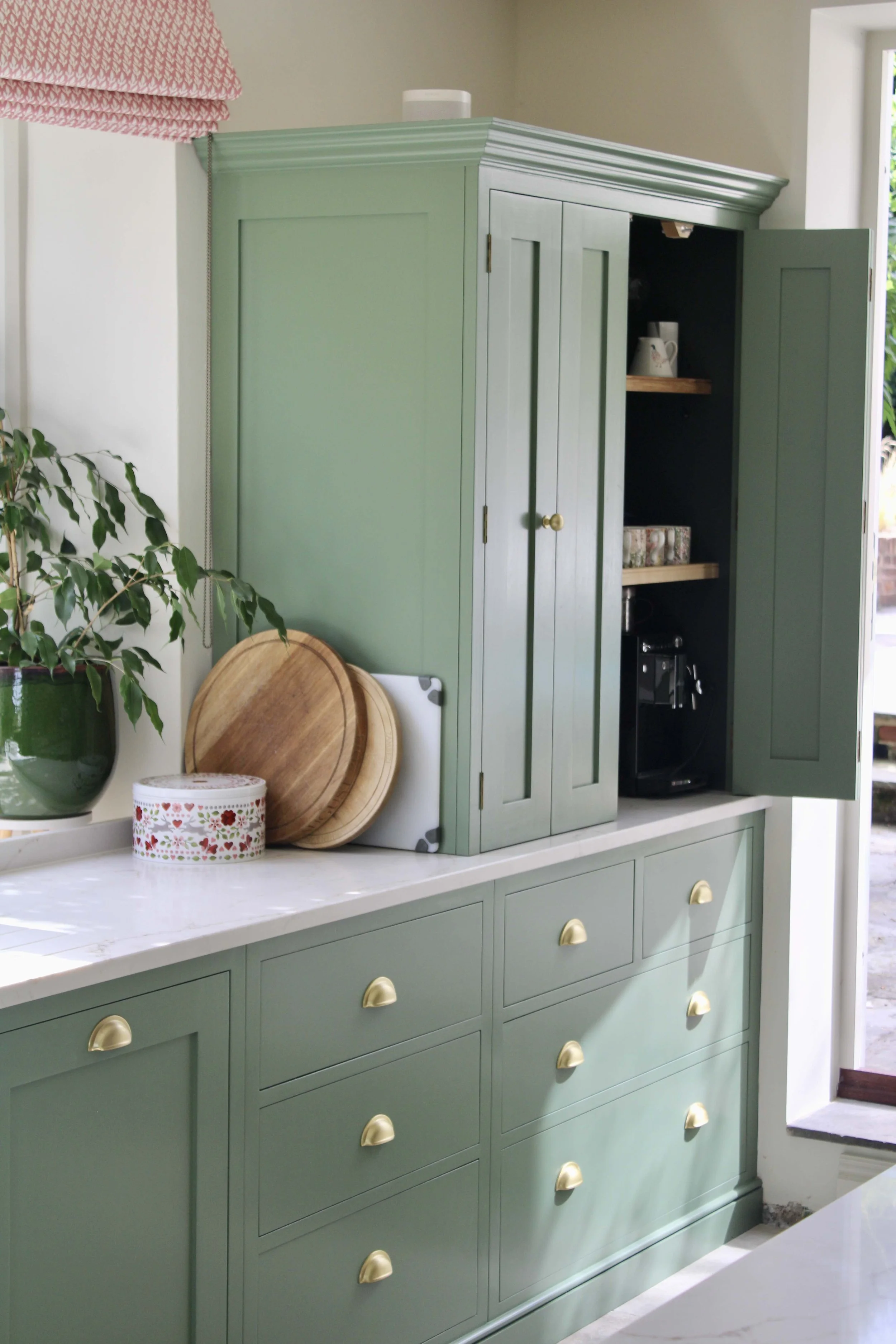 Open kitchen cabinet with two drawers and four shelves, containing small appliances, food items, and kitchenware, in a bright kitchen with wooden floors and a window.