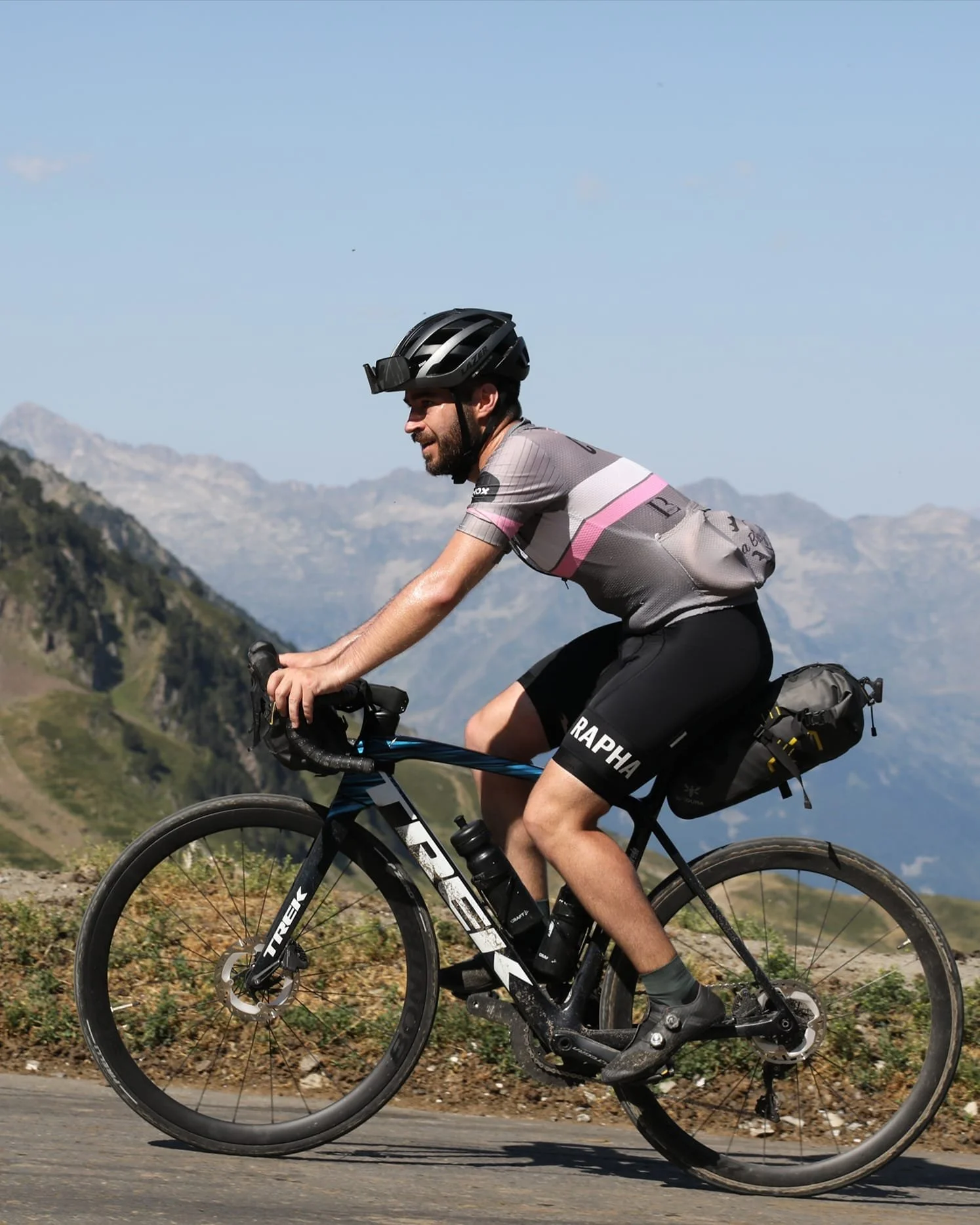 Un homme fait du vélo sur une route de montagne par une journée ensoleillée, portant un casque, une tenue de cycliste, et un sac à dos, avec des montagnes en arrière-plan.