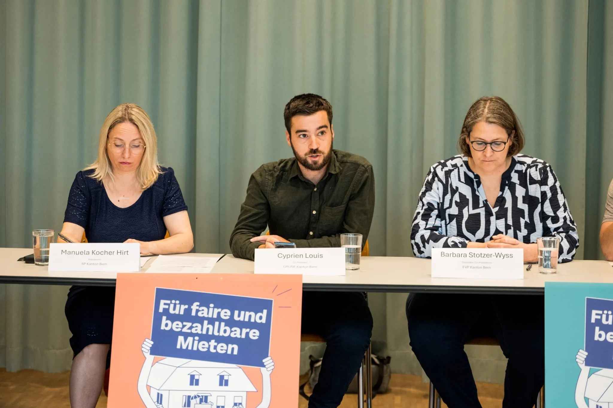 Trois personnes assises à une table lors d'une réunion ou d'une conférence, avec des pancartes mentionnant la lutte contre les loyers abordables.