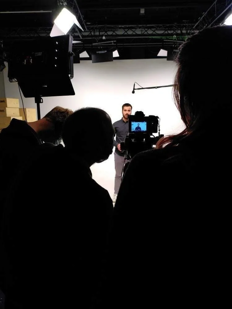 Une personne est en train d'enregistrer une vidéo ou de prendre une photo dans un studio, avec des membres de l'équipe technique autour, au fond un fond blanc, et des lumières et matériel d'enregistrement.
