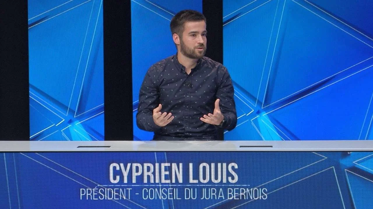 Un homme parle lors d'une émission de télévision avec un fond bleu et une table portant le nom Cyprien Louis, président du Conseil du Jura Bernois.