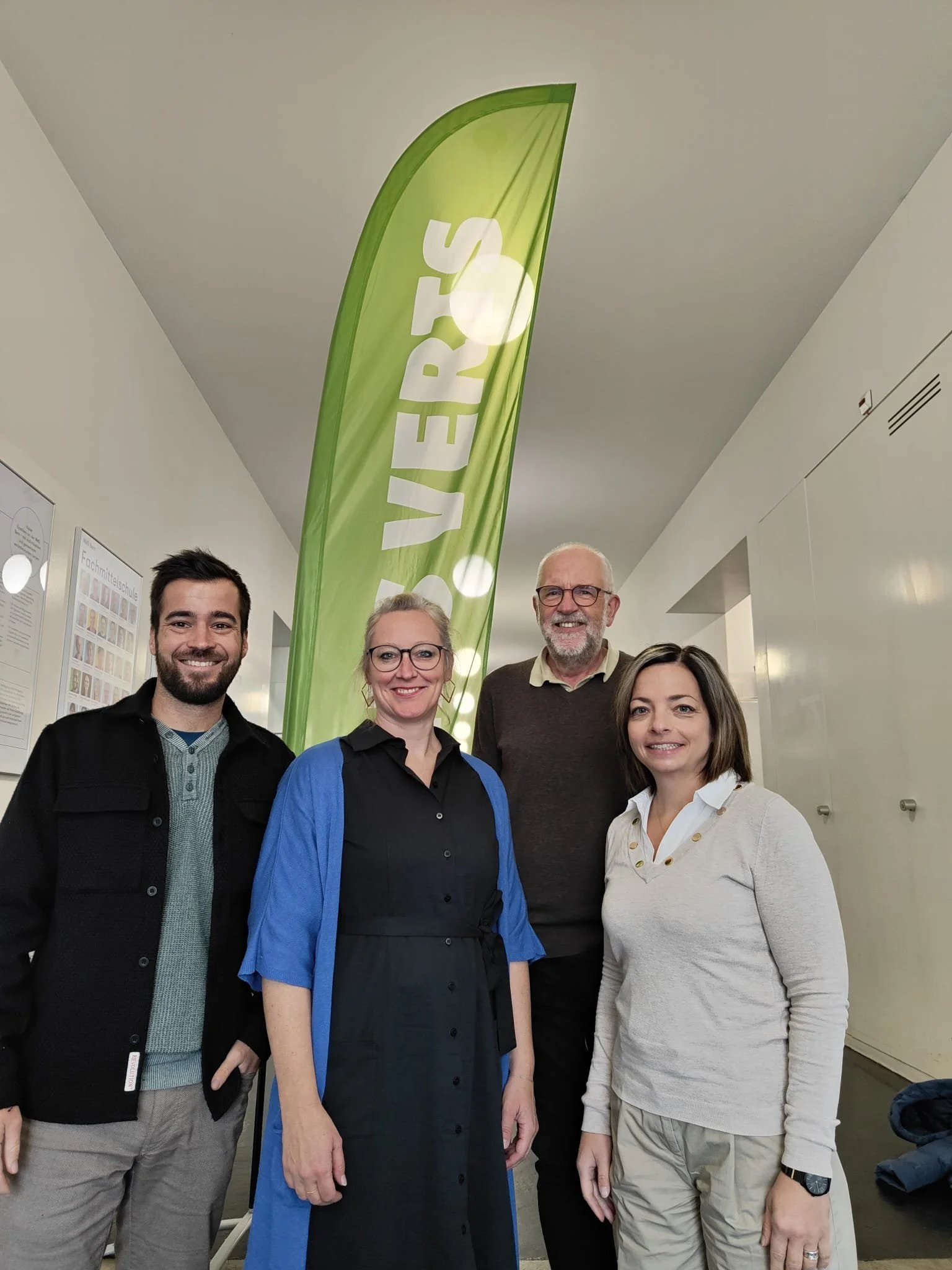Groupe de quatre personnes souriantes posant devant un drapeau vert avec le mot 'VERIS' écrit dessus, dans un intérieur bien éclairé.