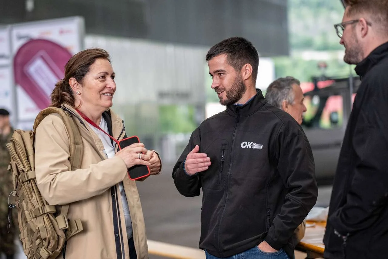 Groupe de trois personnes discutant à l'extérieur, deux hommes et une femme, avec la femme souriante et tenant un téléphone, tous habillés en vêtements décontractés.