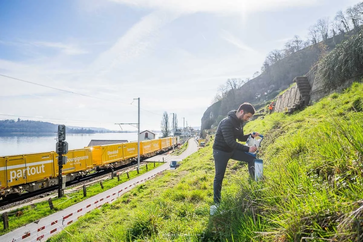 Un homme avec un ordinateur portable en plein air près d'une voie ferrée, avec un train orange passant à côté, sur une pente herbeuse sous un ciel ensoleillé.
