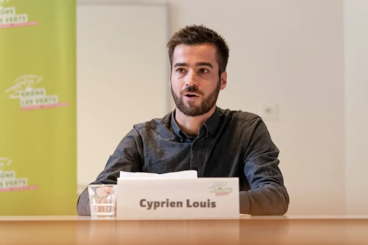 Un homme avec une barbe et des cheveux courts assis à une table, portant une chemise noire. Sur la table, un nom en carton: Cyprien Louis, un verre d'eau. En arrière-plan, un panneau vert avec un logo et du texte en blanc.