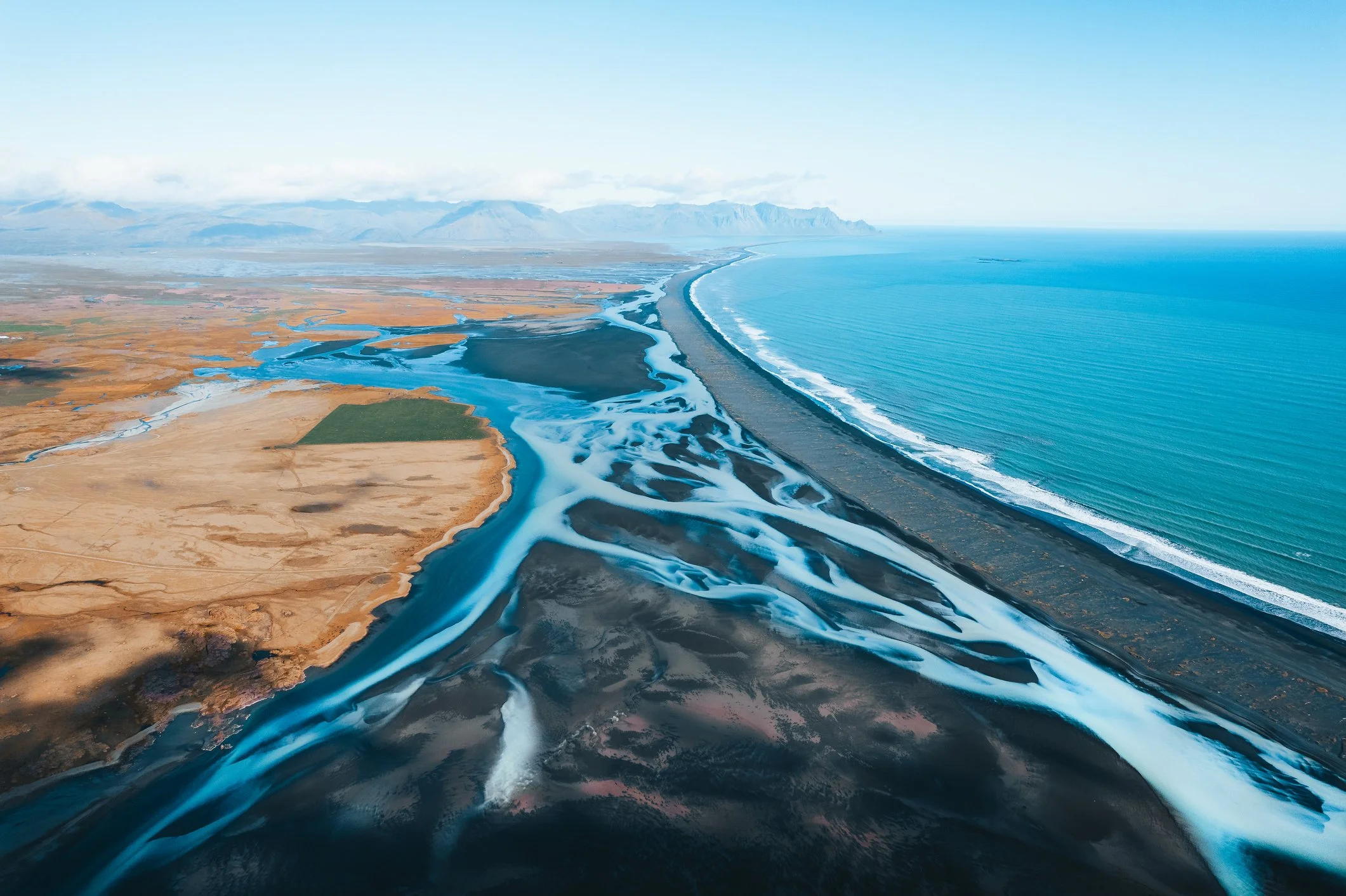 Aerial view of a coastline with a sandy beach, blue ocean waves, and a river flowing into the sea near mountains in the background.