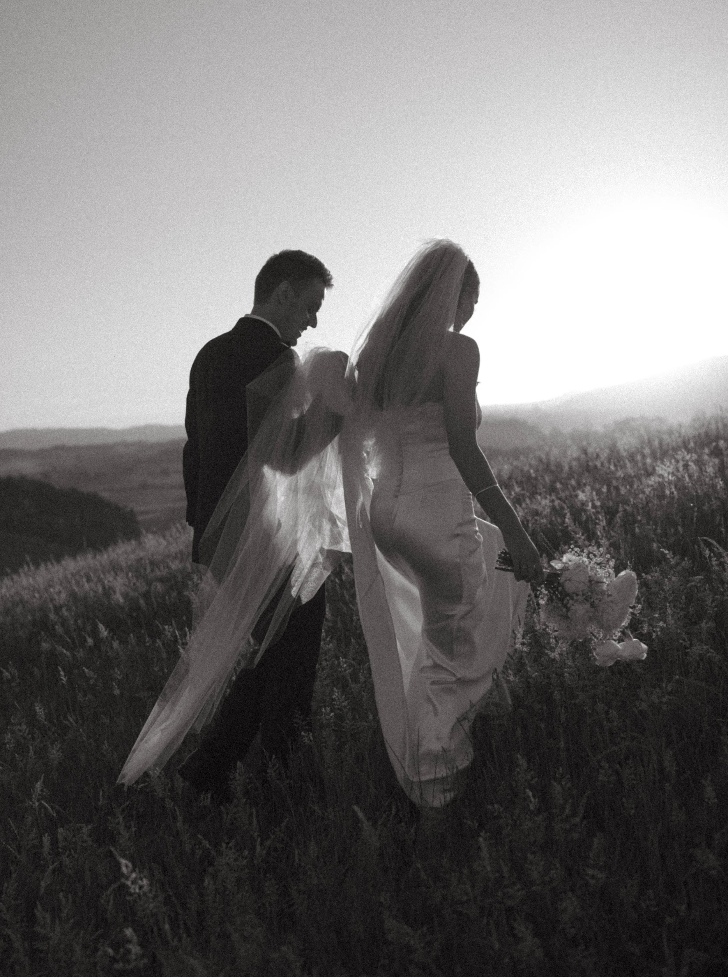A black and white photo of a bride and groom walking through a grassy field at sunset, with the bride holding a bouquet of flowers.