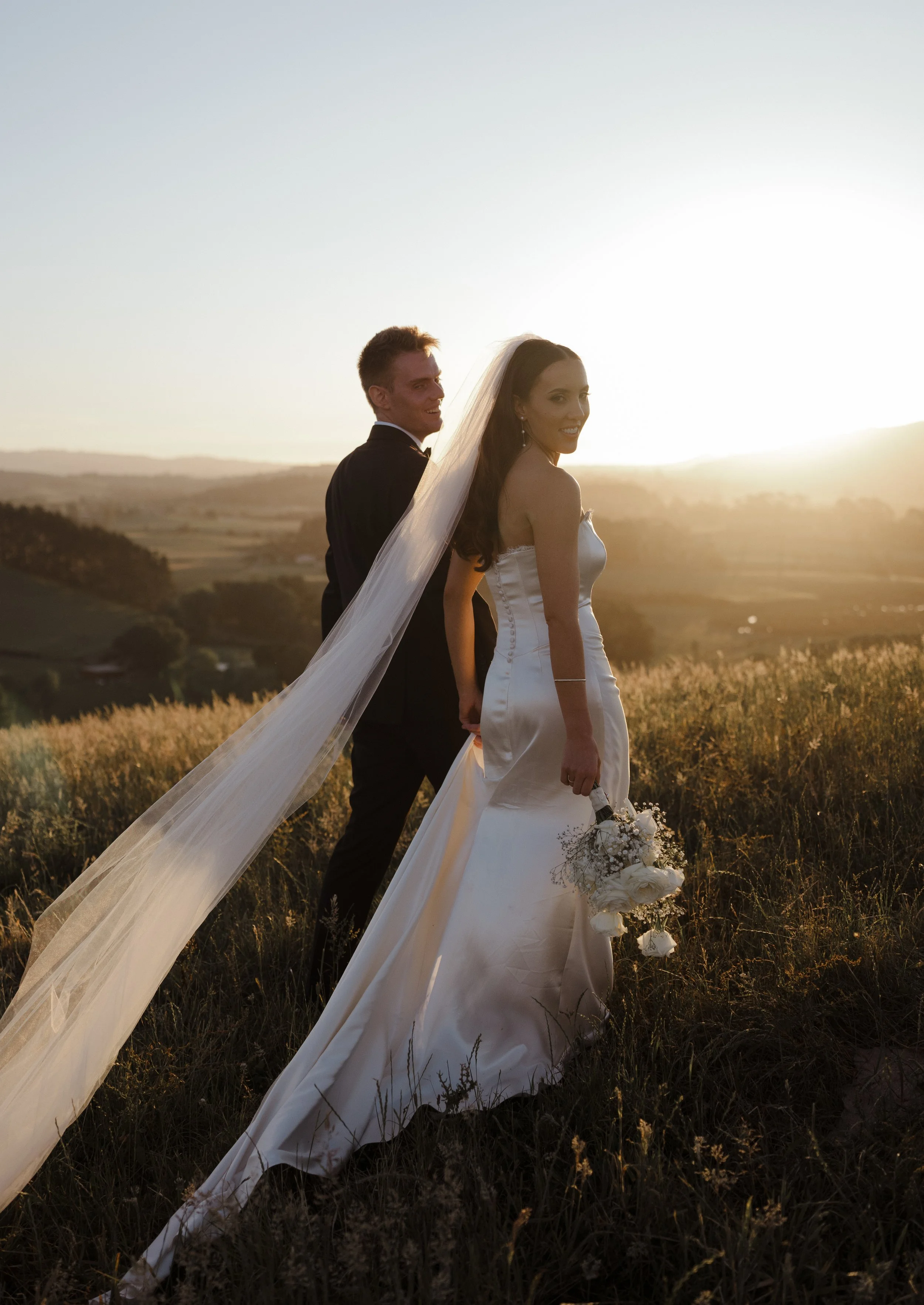 A bride and groom stand outdoors in a field at sunset, holding hands. The bride wears a white wedding gown and veil, holding a bouquet of white flowers. The groom wears a dark tuxedo. The landscape features rolling hills and the sky is illuminated by