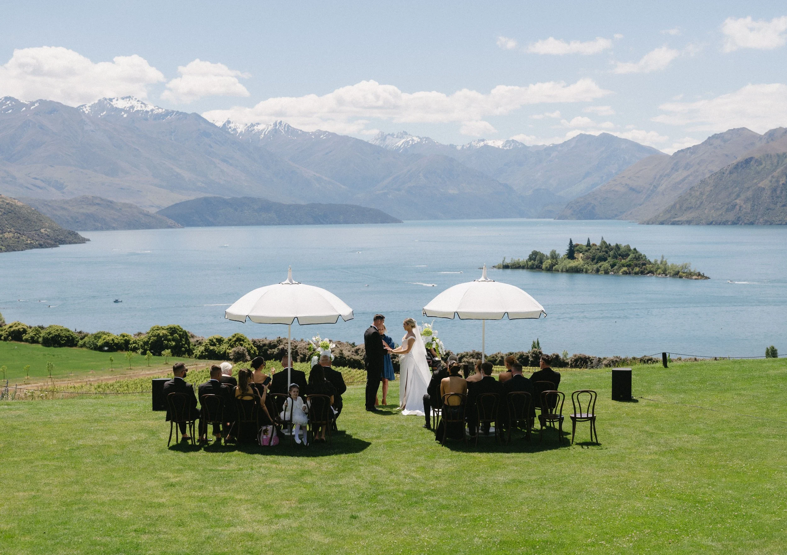 A wedding ceremony taking place outdoors on a grassy area near a lake, with mountains in the background. The bride and groom are exchanging vows under a white umbrella, with guests seated around them and three other umbrellas providing shade.