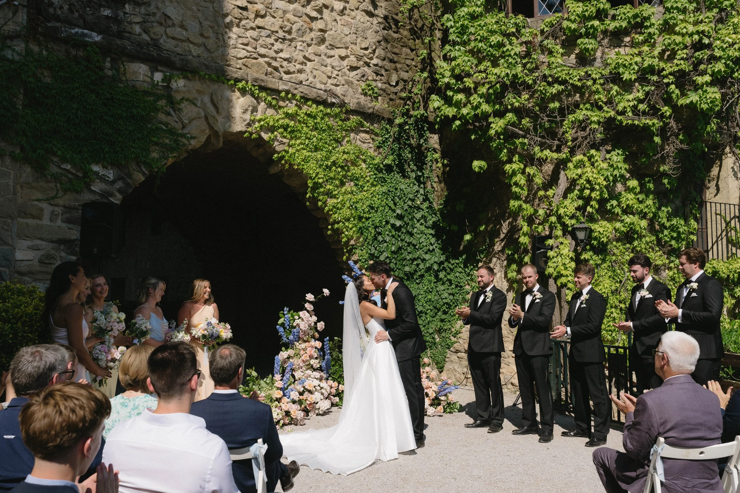 A wedding ceremony taking place outdoors under a stone archway covered with green vines. A bride and groom are exchanging vows, with the bride in a white gown and veil, and the groom in a black suit. Bridesmaids and groomsmen are standing nearby, and