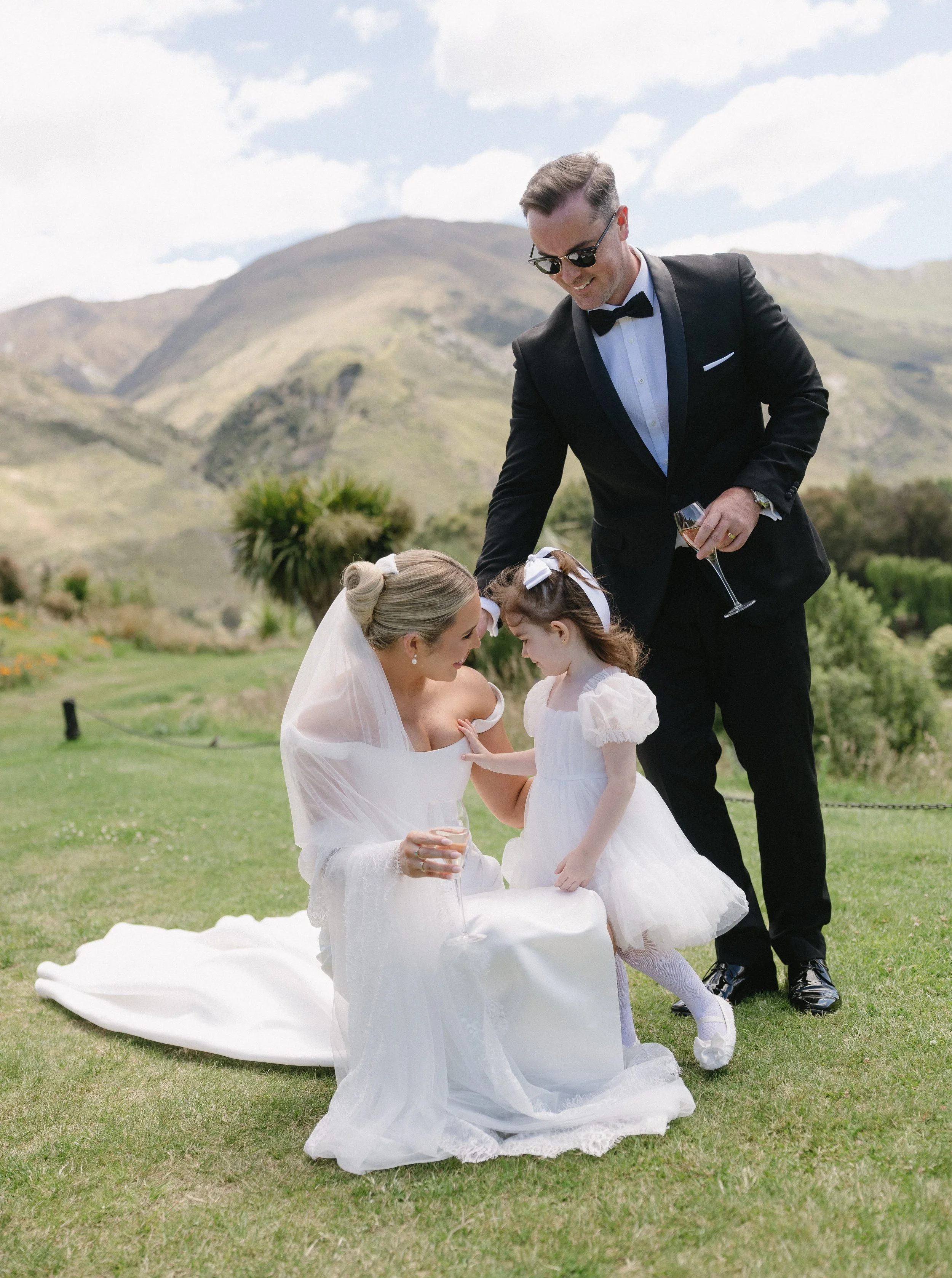 A bride in a white wedding gown and veil, a groom in a black tuxedo with sunglasses, and a young girl in a white dress interacting outdoors in a scenic green mountain setting, with the groom holding a glass of champagne.