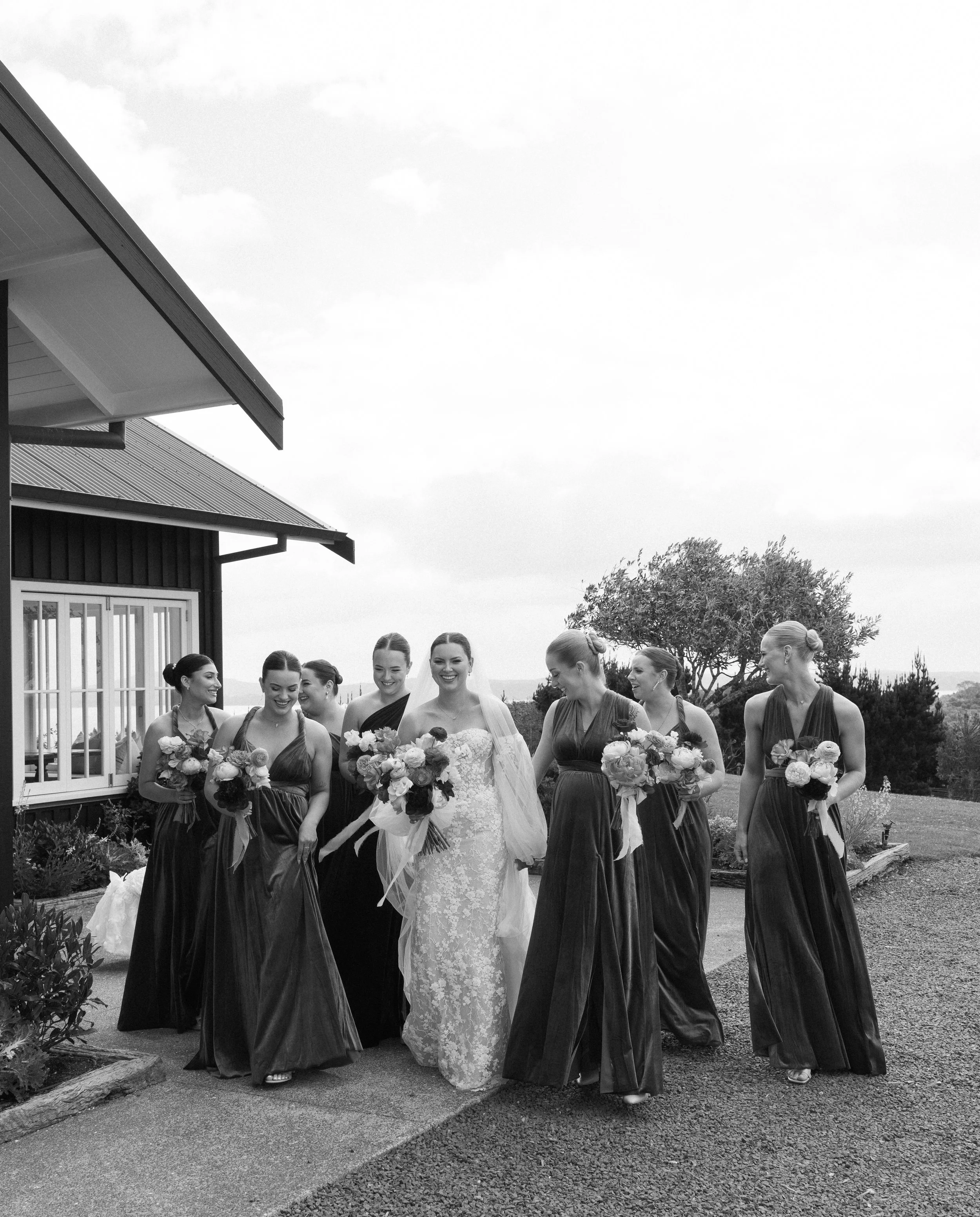 Bride in wedding dress walking with bridesmaids in long dresses holding bouquets outside near house and trees.