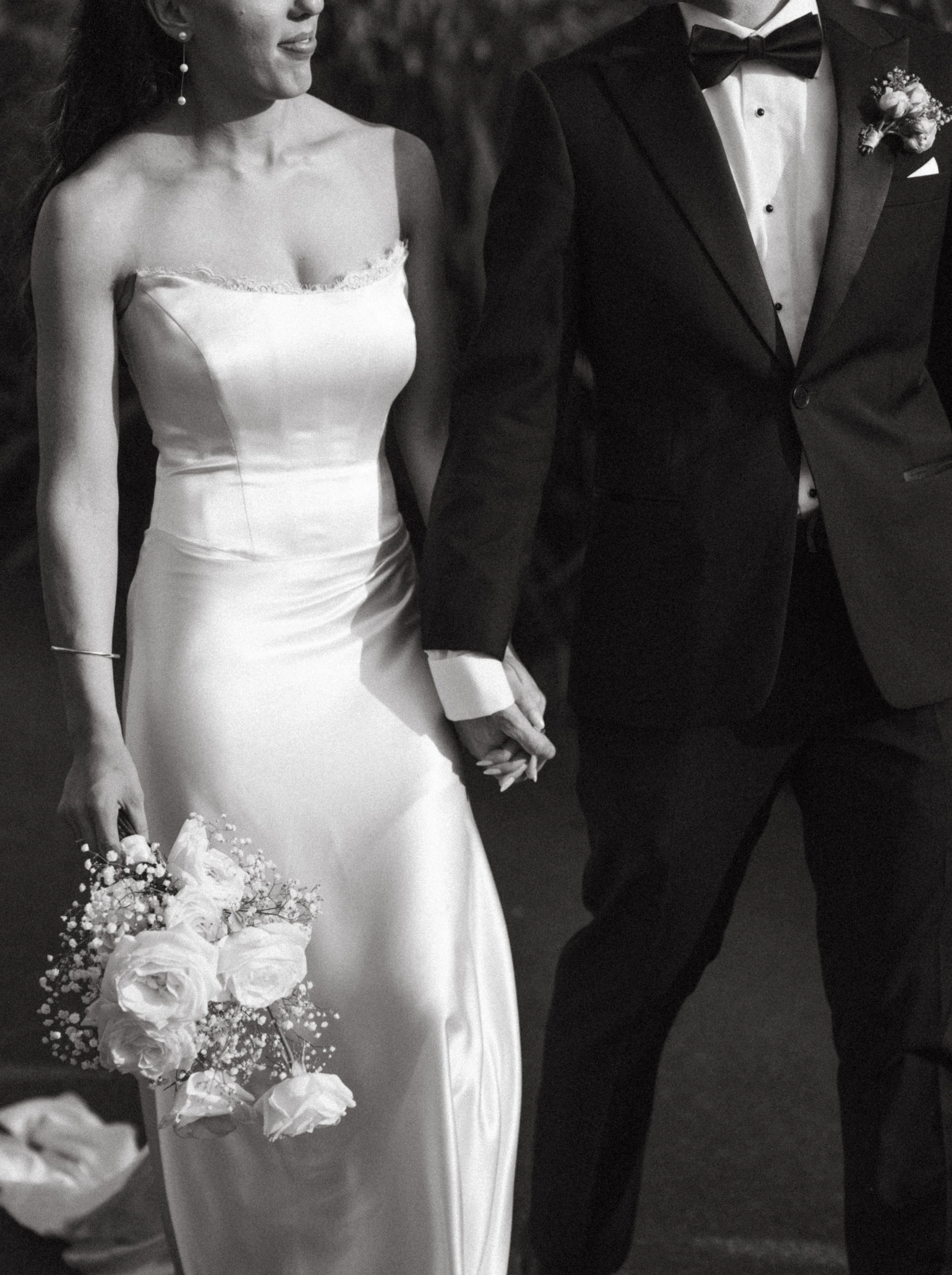 A bride and groom holding hands, the bride in a strapless wedding gown holding a bouquet of flowers, and the groom in a tuxedo with a boutonniere.